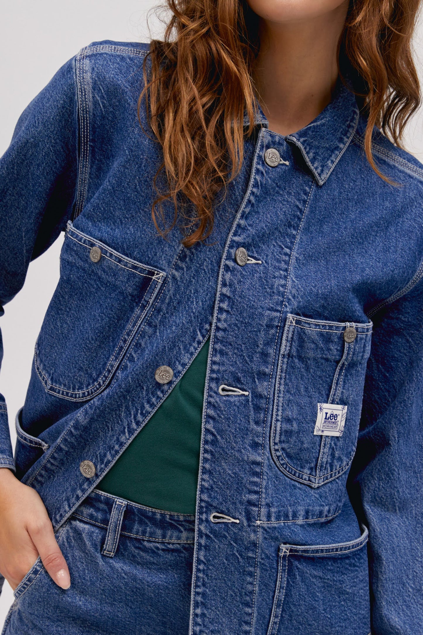 A curly-haired model featuring a blue chore jacket designed with triple-stitched hems and roomy pockets on an oversized fit.