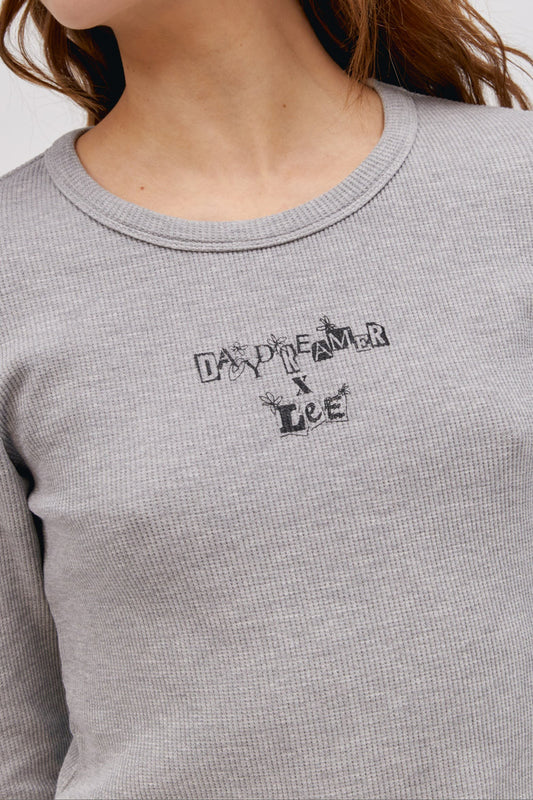 A curly-haired model featuring a heather grey colored shrunken thermal made with a slim fit, a cropped length, and designed with 'Daydreamer x Lee' in an original cut - paste style treatment accented with hand drawn graphic details.