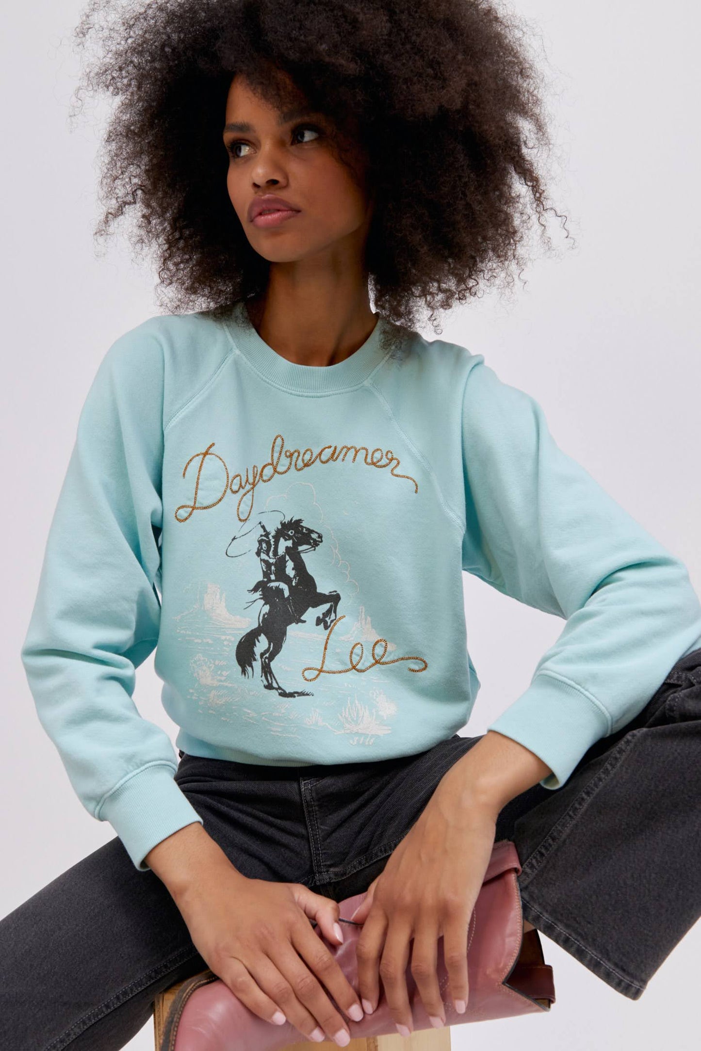 A curly-haired model featuring a raglan crew in icy moon with a bucking cowgirl graphic inspired by the spirit of the West lands on a lightweight, french terry raglan crew embroidered with lasso lettering.