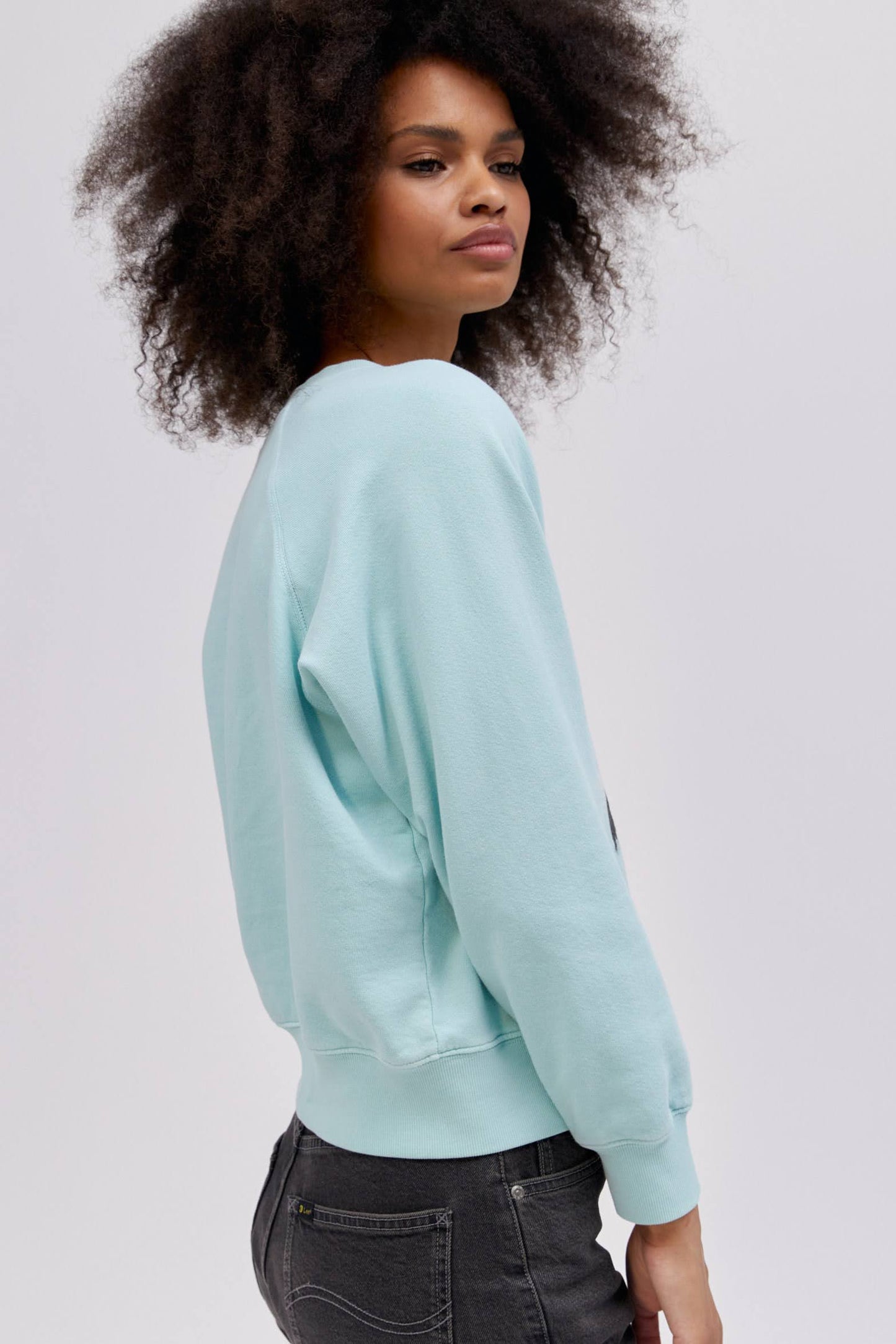 A curly-haired model featuring a raglan crew in icy moon with a bucking cowgirl graphic inspired by the spirit of the West lands on a lightweight, french terry raglan crew embroidered with lasso lettering.