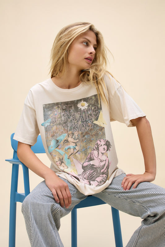 A model featuring a white merch tee stamped with 'The Smashing Pumpkins' and a bunch of angels, statues, and flowers around.