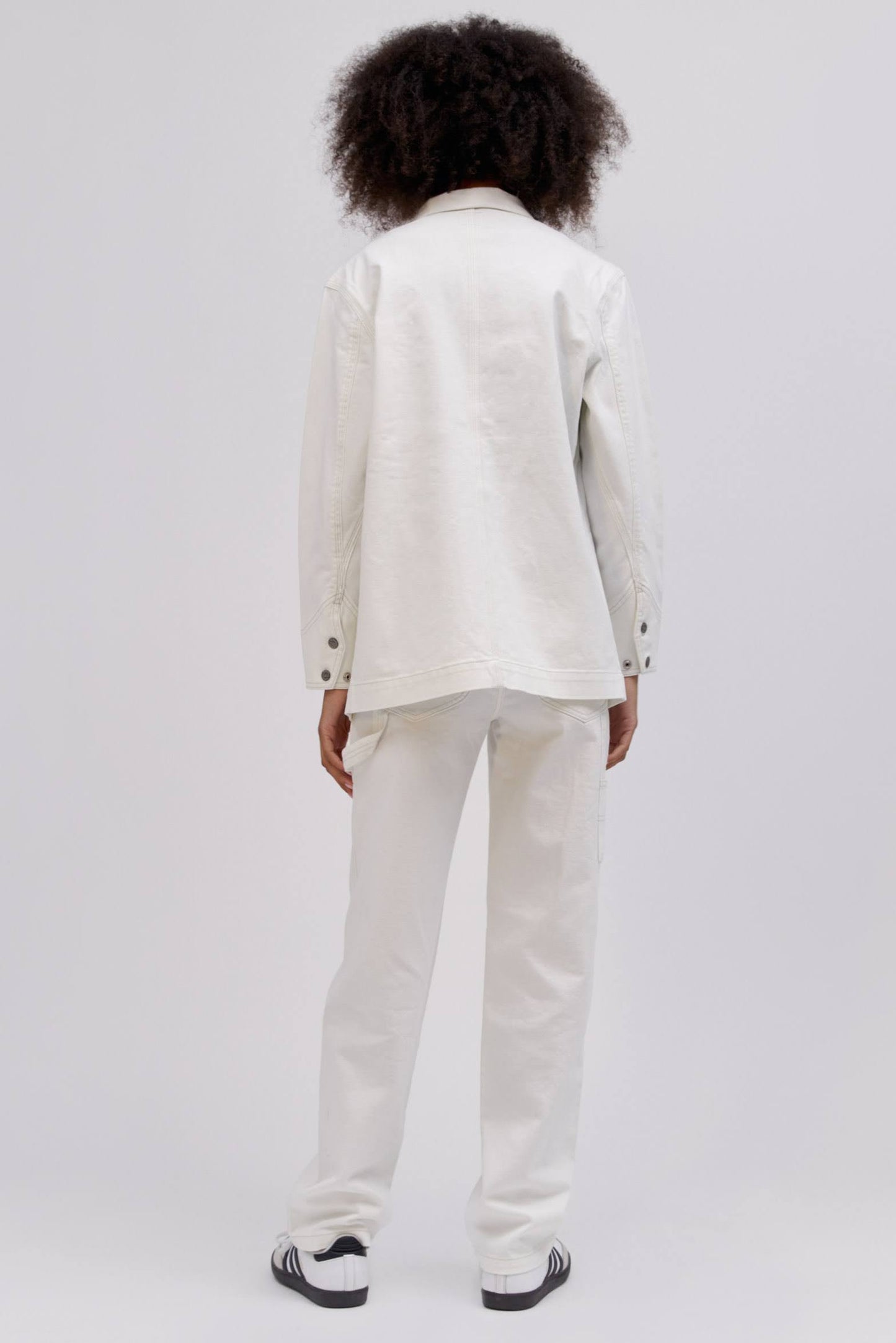 A curly-haired model featuring a white chore jacket designed with  triple-stitched hems and roomy pockets on an oversized fit.