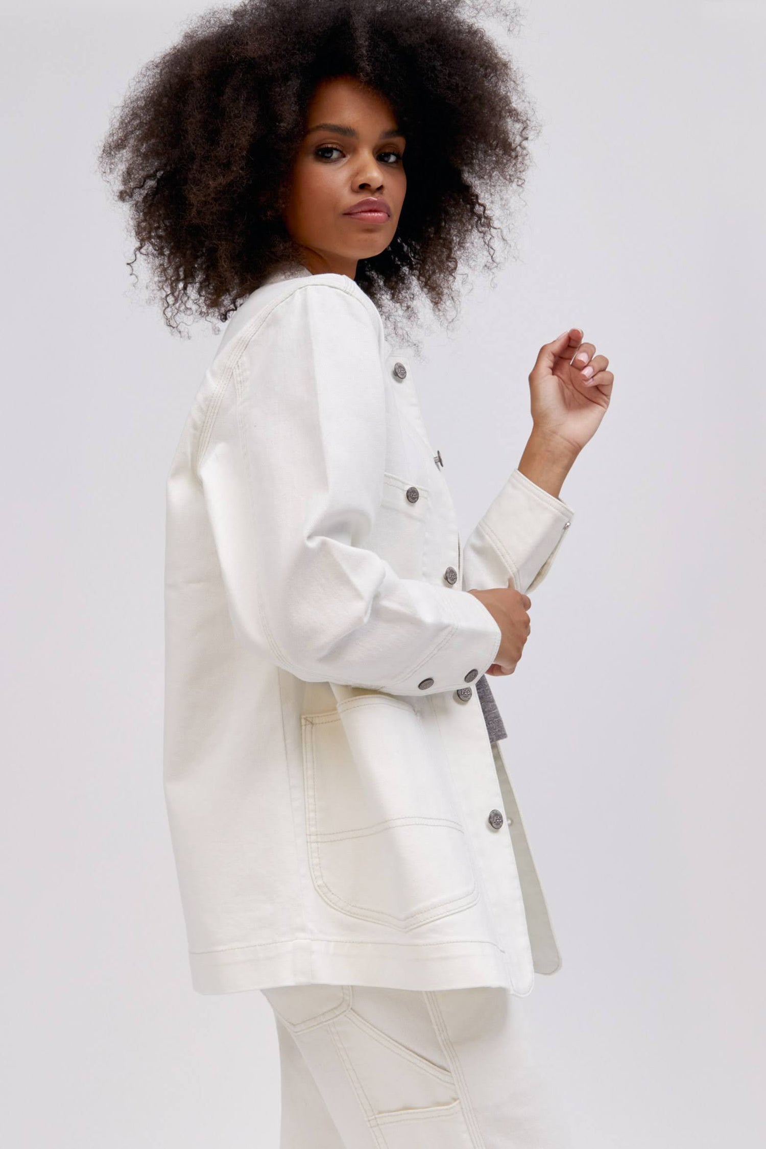 A curly-haired model featuring a white chore jacket designed with  triple-stitched hems and roomy pockets on an oversized fit.