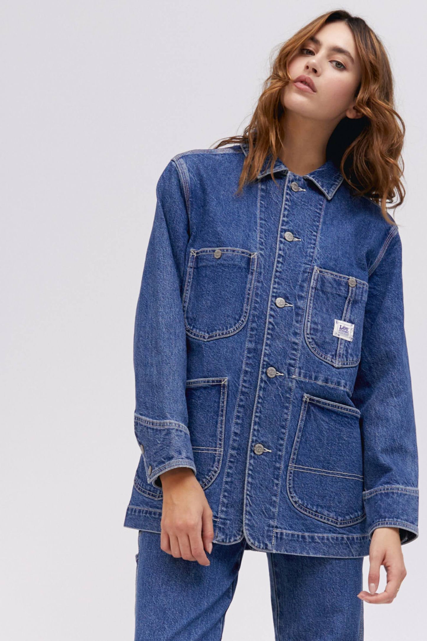 A curly-haired model featuring a blue chore jacket designed with  triple-stitched hems and roomy pockets on an oversized fit.