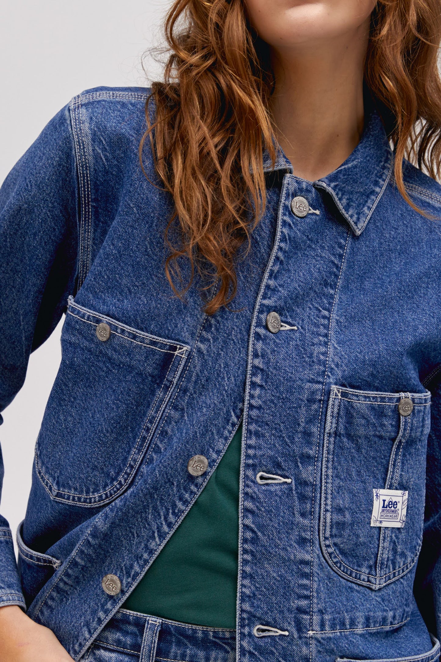 A curly-haired model featuring a blue chore jacket designed with  triple-stitched hems and roomy pockets on an oversized fit.
