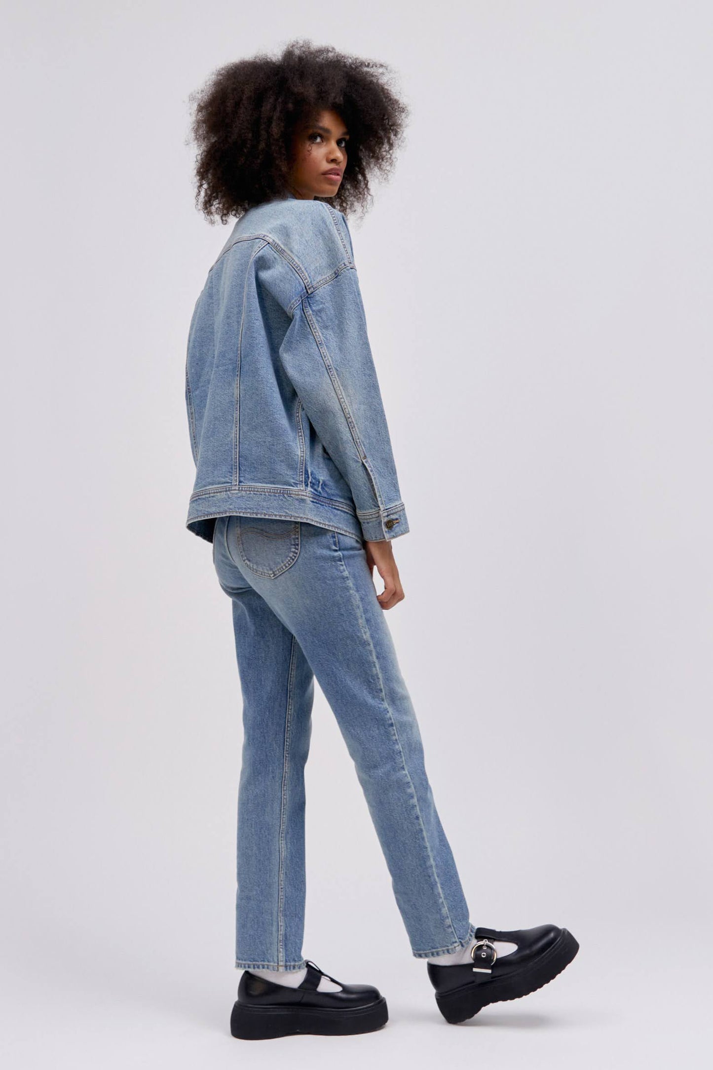 Backside posing curly haired model wearing a reimagined Lee Denim rider jacket in an oversized fit.
