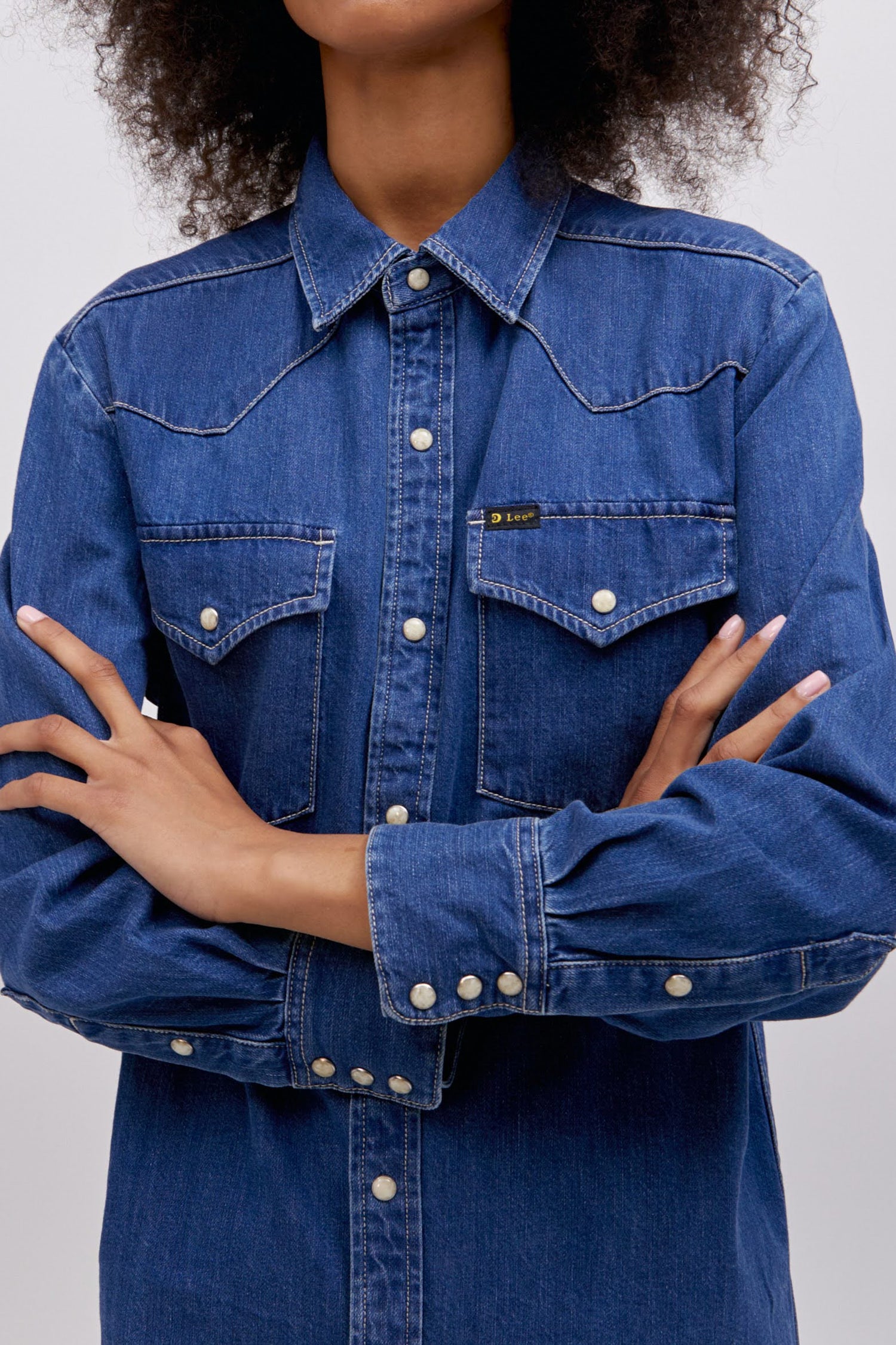 A curly-haired model featuring a timeless blue Western Shirt with a pointed front yoke, chest pockets, and a set of pearl snaps down the center. 