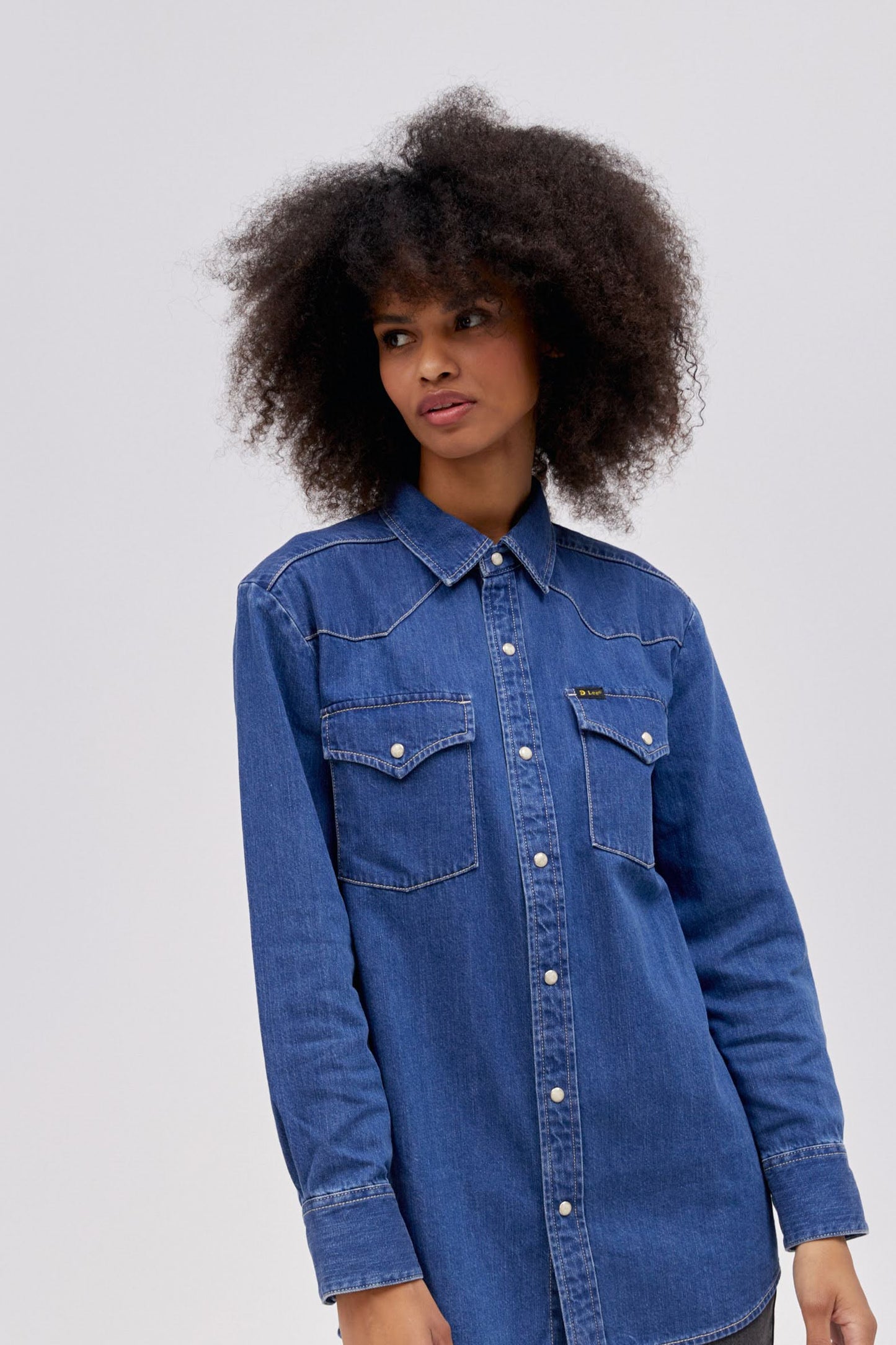 A curly-haired model featuring a timeless blue Western Shirt with a pointed front yoke, chest pockets, and a set of pearl snaps down the center. 