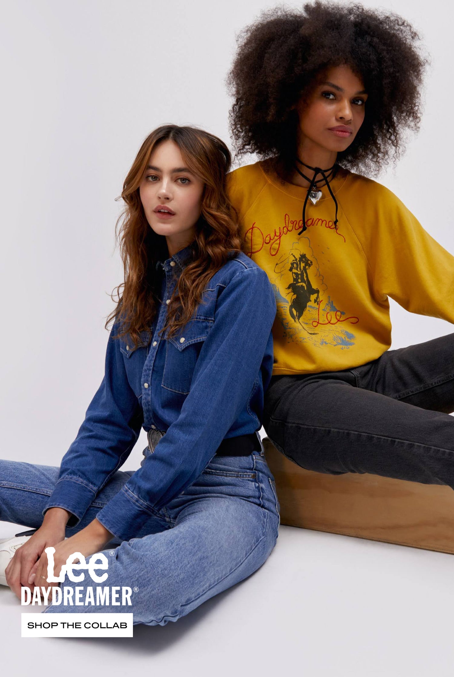 Brunette model wearing denim button up shirt and jeans, curly hair model wearing mustard crewneck, both sitting