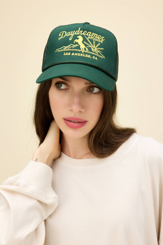 Model wearing a dark green trucker hat with western-style 'Daydreamer Studios' embroidery on the front.