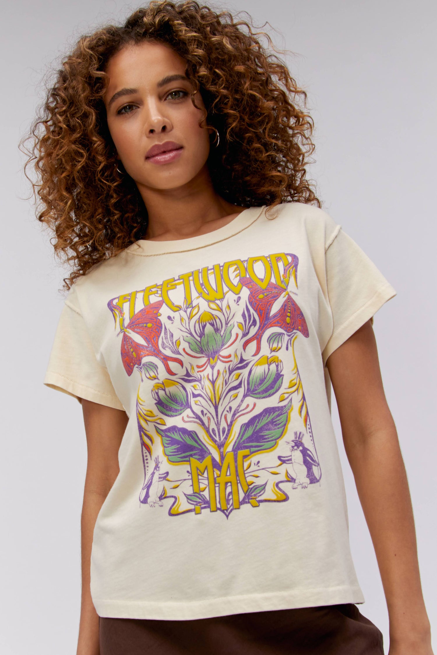 A curly-haired model featuring a parchment-colored tee stamped with the band's name, designed with a graphic of leaves, flowers, and butterflies on the center.