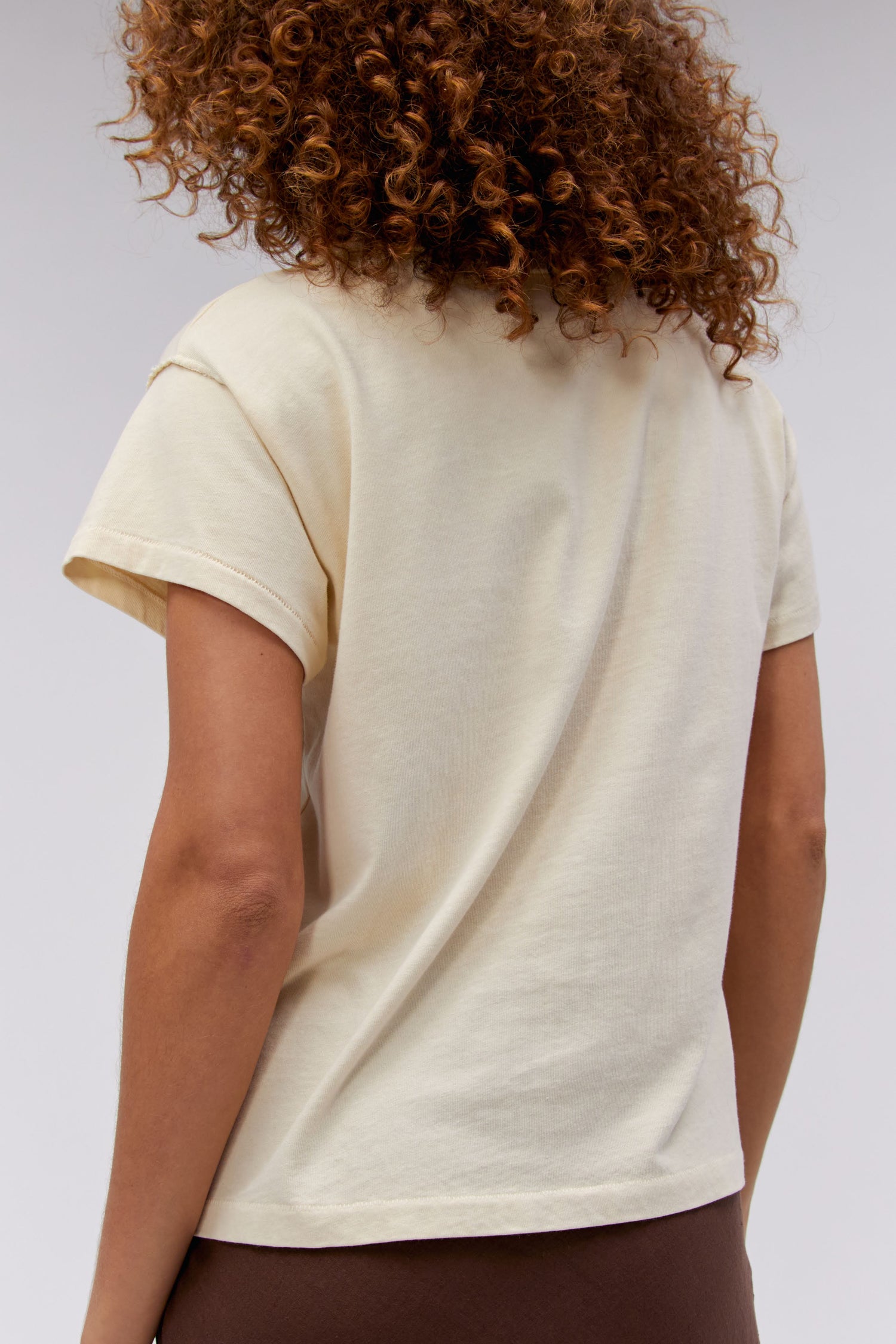 A curly-haired model featuring a parchment-colored tee stamped with the band's name, designed with a graphic of leaves, flowers, and butterflies on the center.
