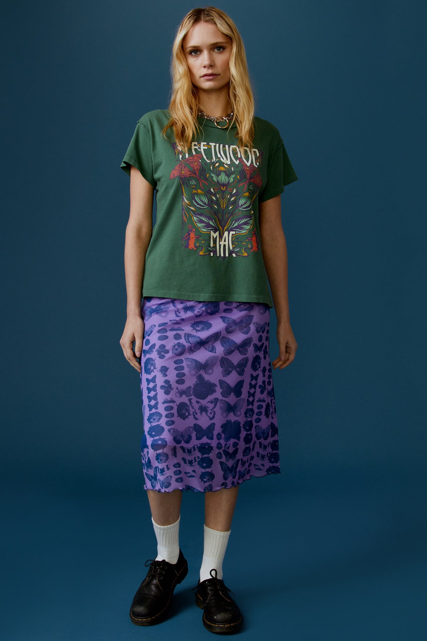 A blonde-haired model featuring a stormy green tee stamped with the band's name, designed with a graphic of leaves, flowers, and butterflies on the center.