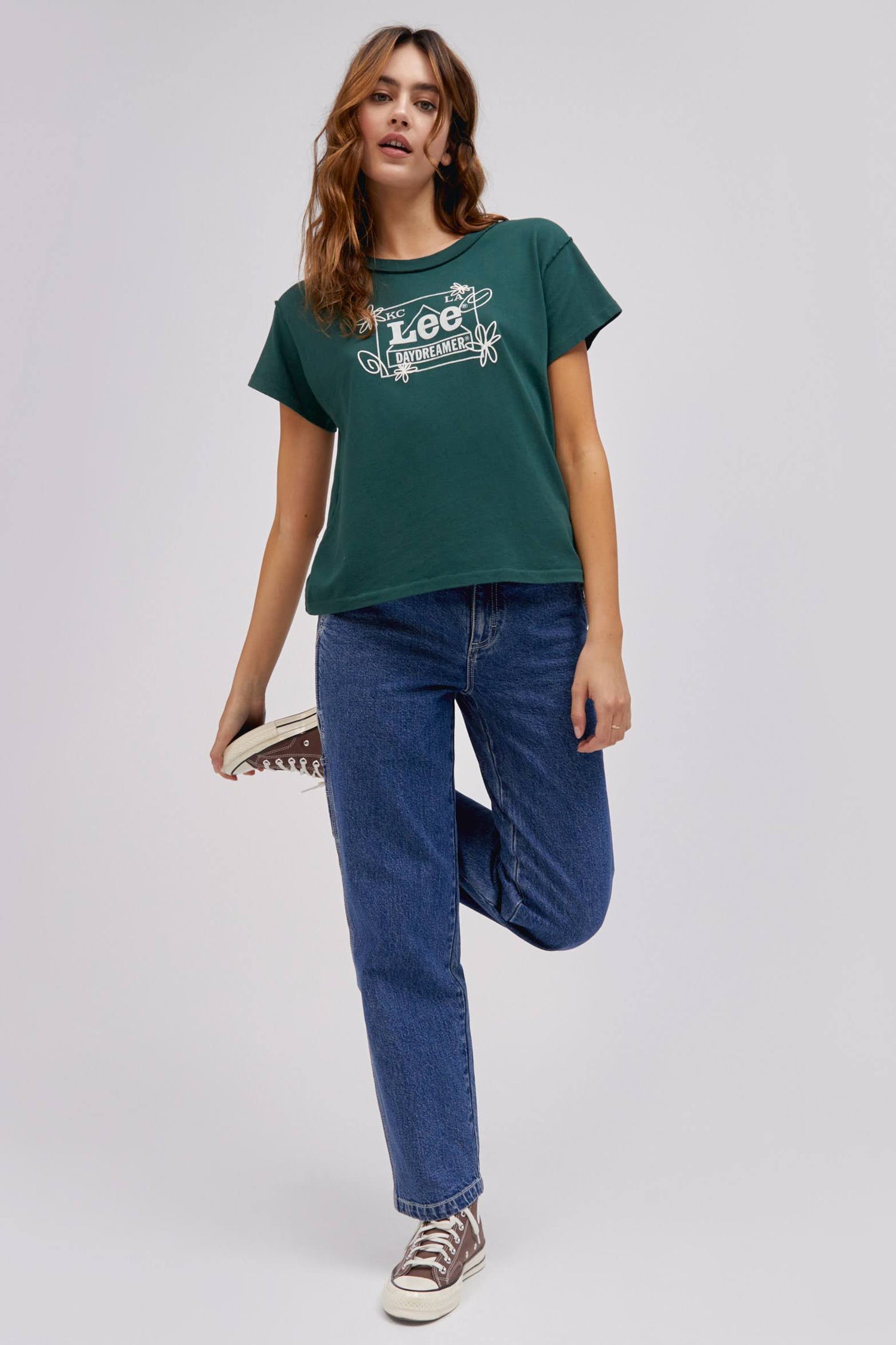 A curly-haired model featuring a pine reverse gf tee designed with the original Lee workwear logo reimagined into an exclusive, co-branded graphic with layered doodles in puff ink.