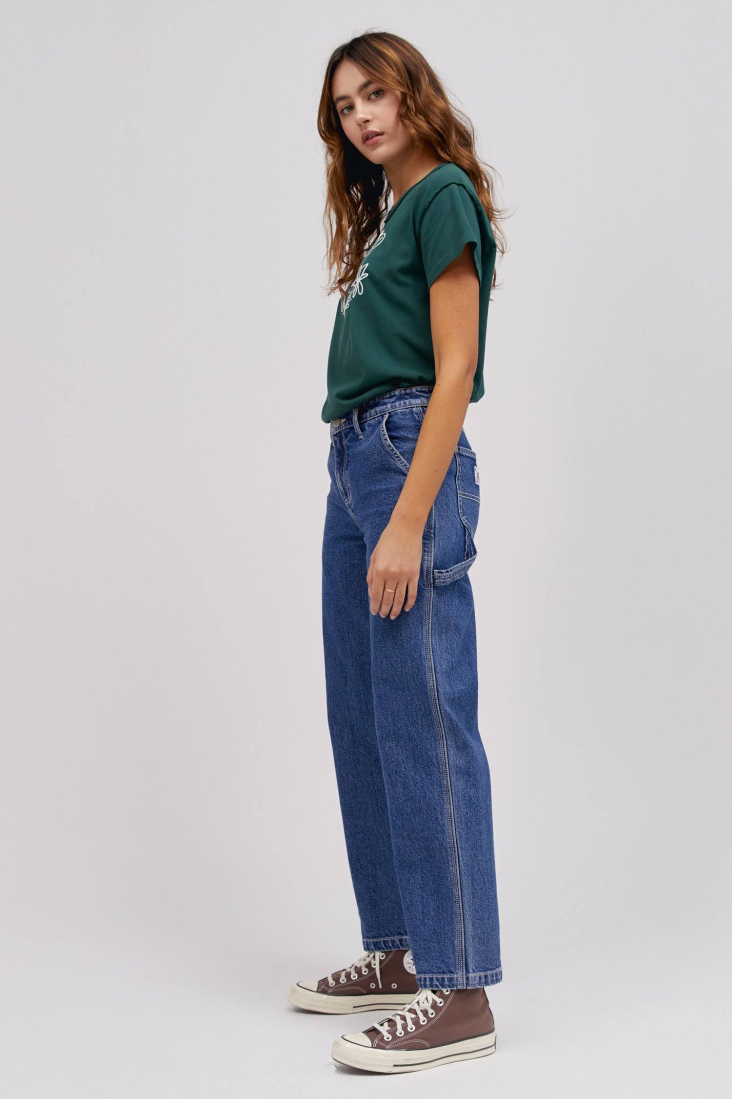 A curly-haired model featuring a pine reverse gf tee with a blue workwear pant made from heavy cotton, accented with key Lee detailing of triple-stitched seams, oversized pockets and a uniform like hammer loop.