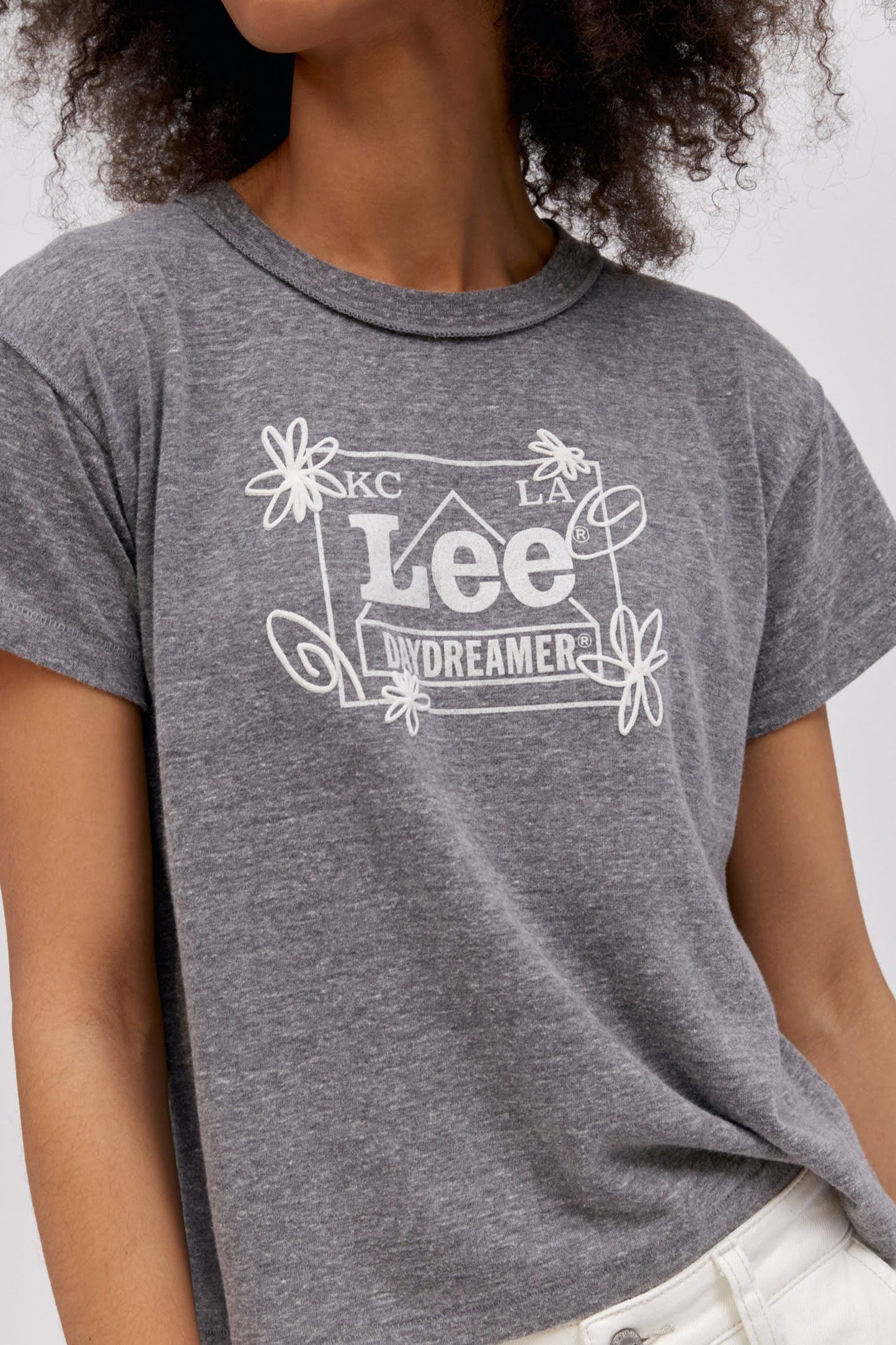 A curly-haired model featuring a heather grey reverse gf tee designed with the original Lee workwear logo reimagined into an exclusive, co-branded graphic with layered doodles in puff ink.