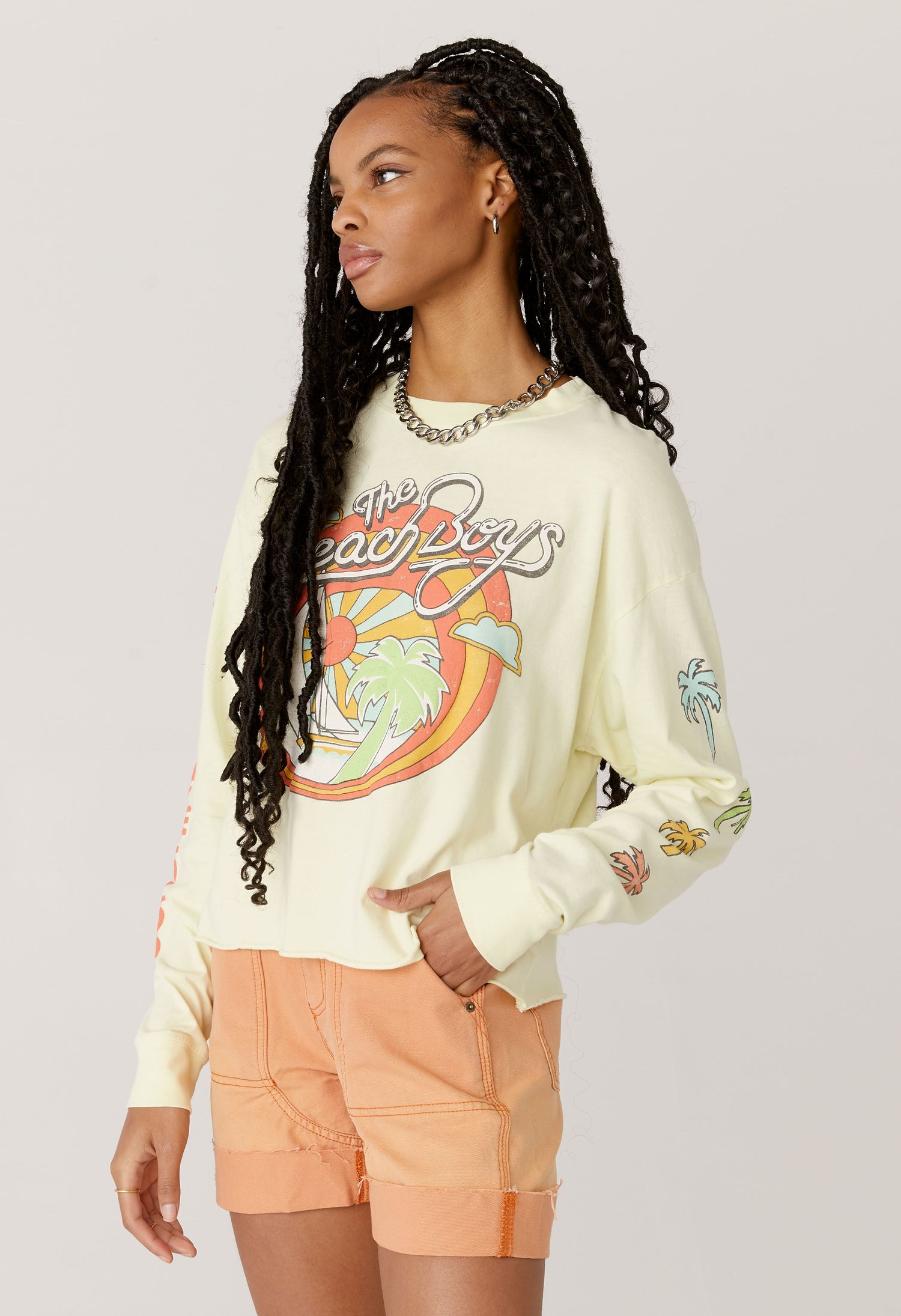 A dark curly haired model featuring a white long sleeve crop designed with the group’s name, iconic summertime font and graphics. 