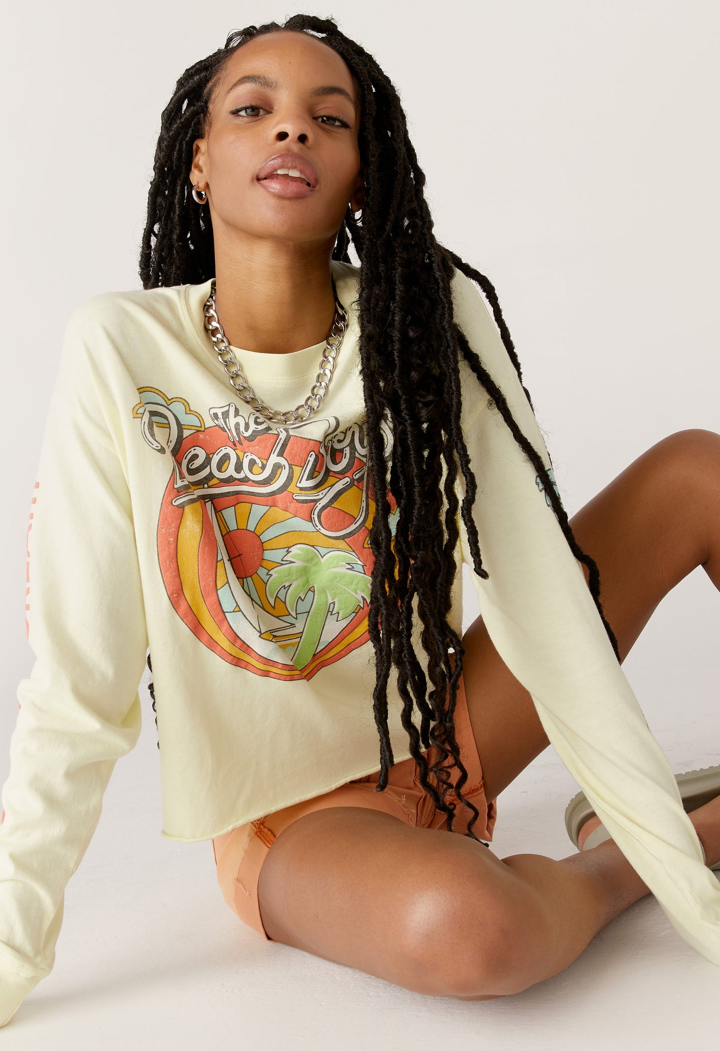 A dark curly haired model featuring a white long sleeve crop designed with the group’s name, iconic summertime font and graphics. 