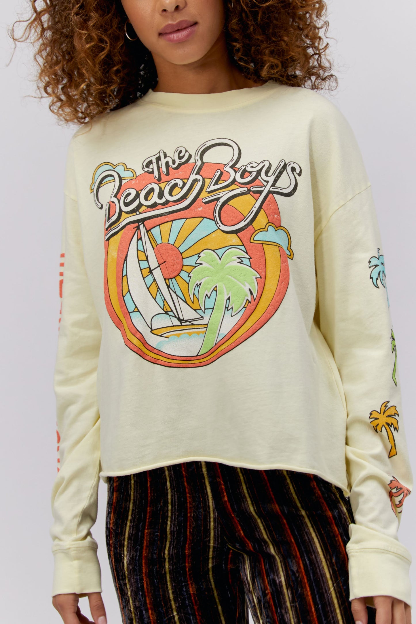 A blonde curly haired model featuring a white long sleeve crop designed with the group’s name, iconic summertime font and graphics. 