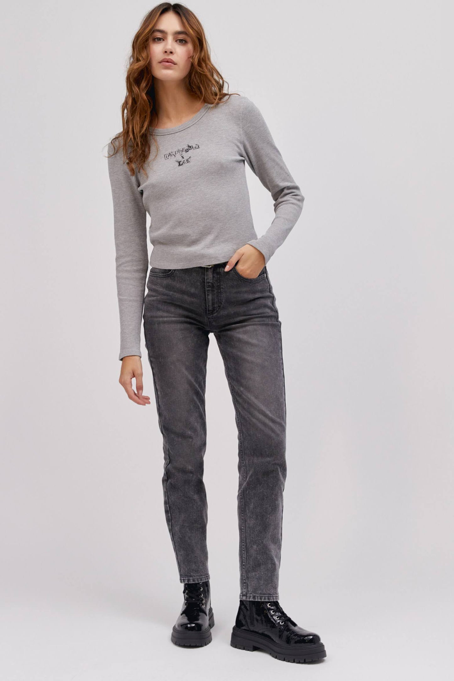 A curly-haired model featuring a heather grey colored shrunken thermal made with a slim fit, a cropped length, and designed with 'Daydreamer x Lee' in an original cut - paste style treatment accented with hand drawn graphic details.