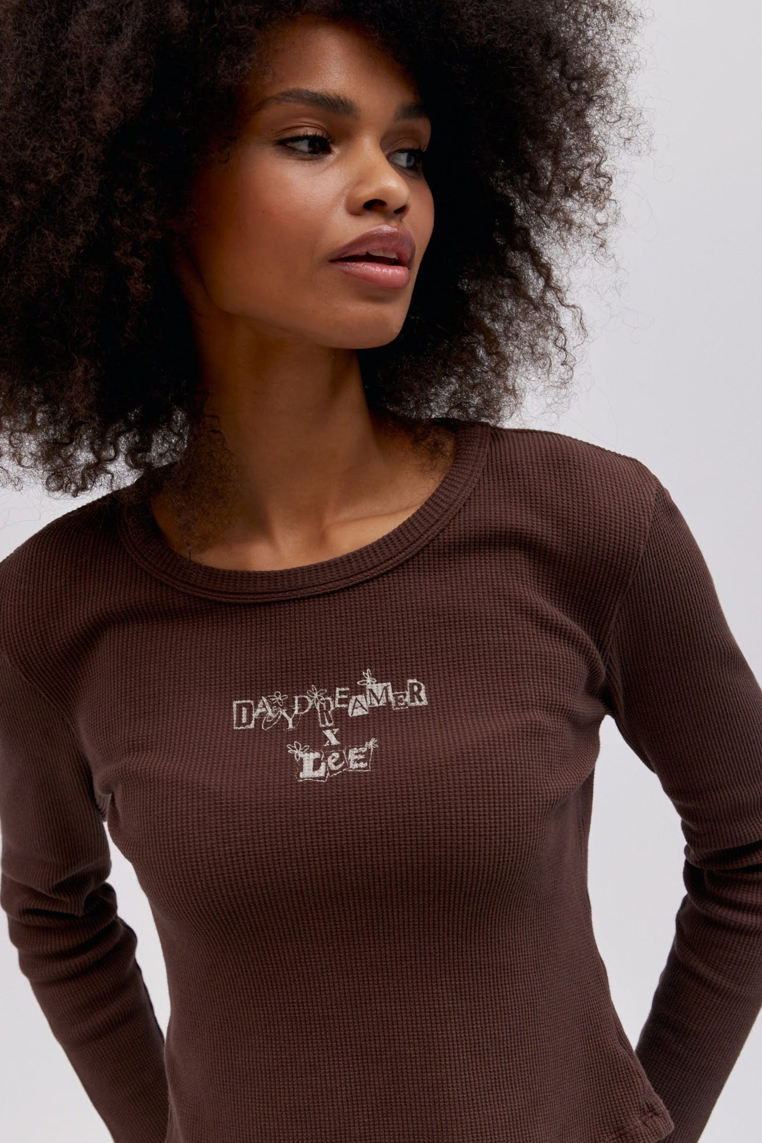 A curly-haired model featuring a coffee quarts colored shrunken thermal made with a slim fit, a cropped length, and designed with 'Daydreamer x Lee' in an original cut - paste style treatment accented with hand drawn graphic details.
