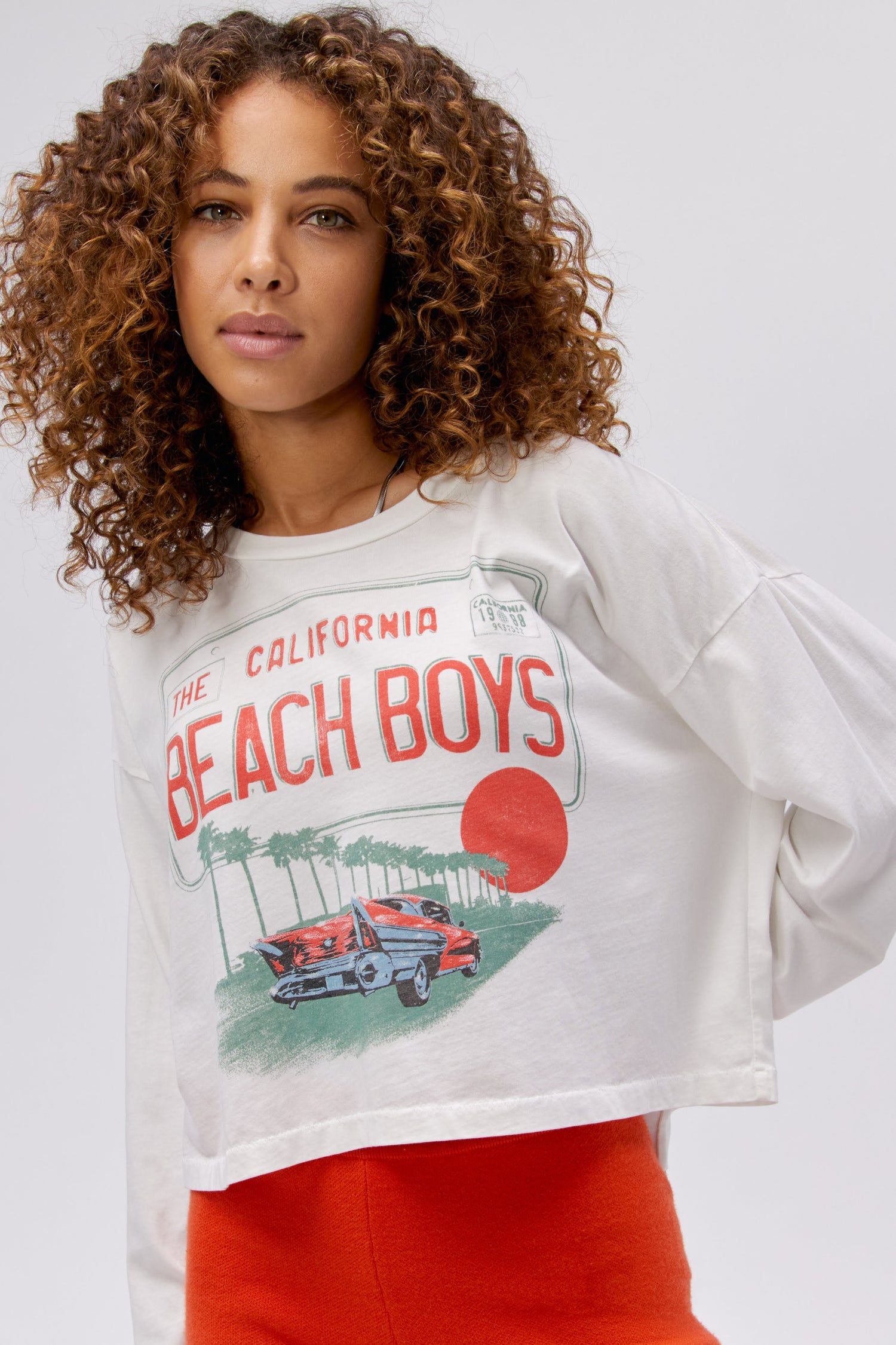 A curly-haired model featuring a white long sleeve stamped with "The Californa Beach Boys" and is desiged with a graphic of trees and cars on the center.