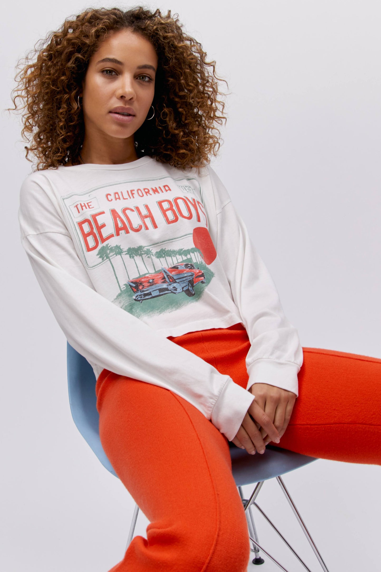 A curly-haired model featuring a white long sleeve stamped with "The Californa Beach Boys" and is desiged with a graphic of trees and cars on the center.