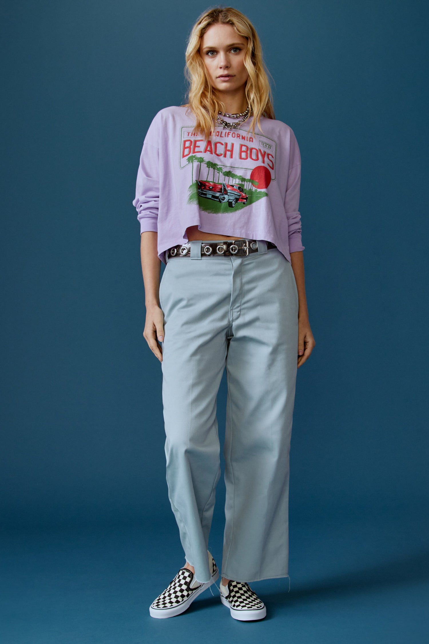 A blonde-haired model featuring a lavender long sleeve stamped with "The Californa Beach Boys" and is desiged with a graphic of trees and cars on the center.