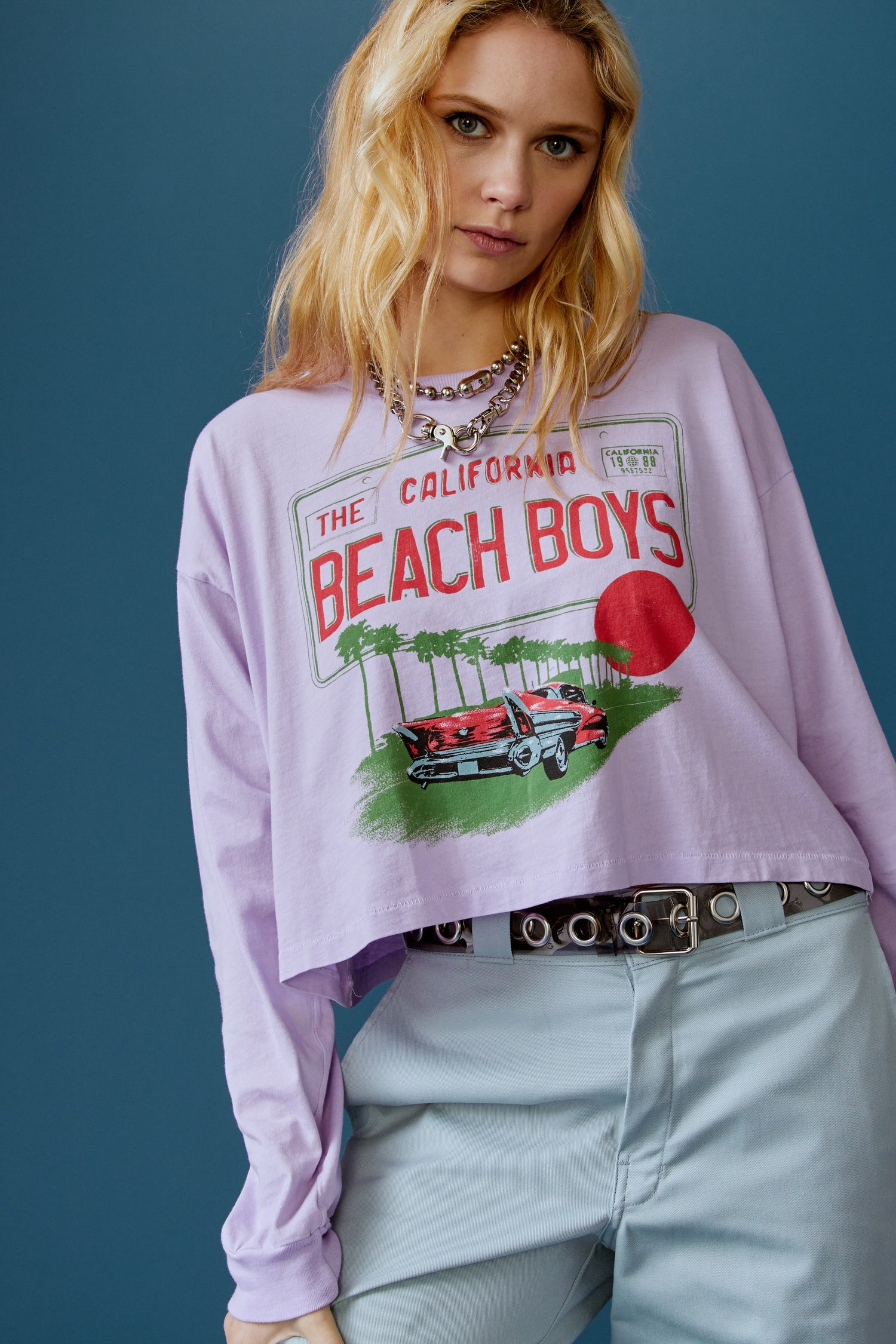 A blonde-haired model featuring a lavender long sleeve stamped with "The Californa Beach Boys" and is desiged with a graphic of trees and cars on the center.