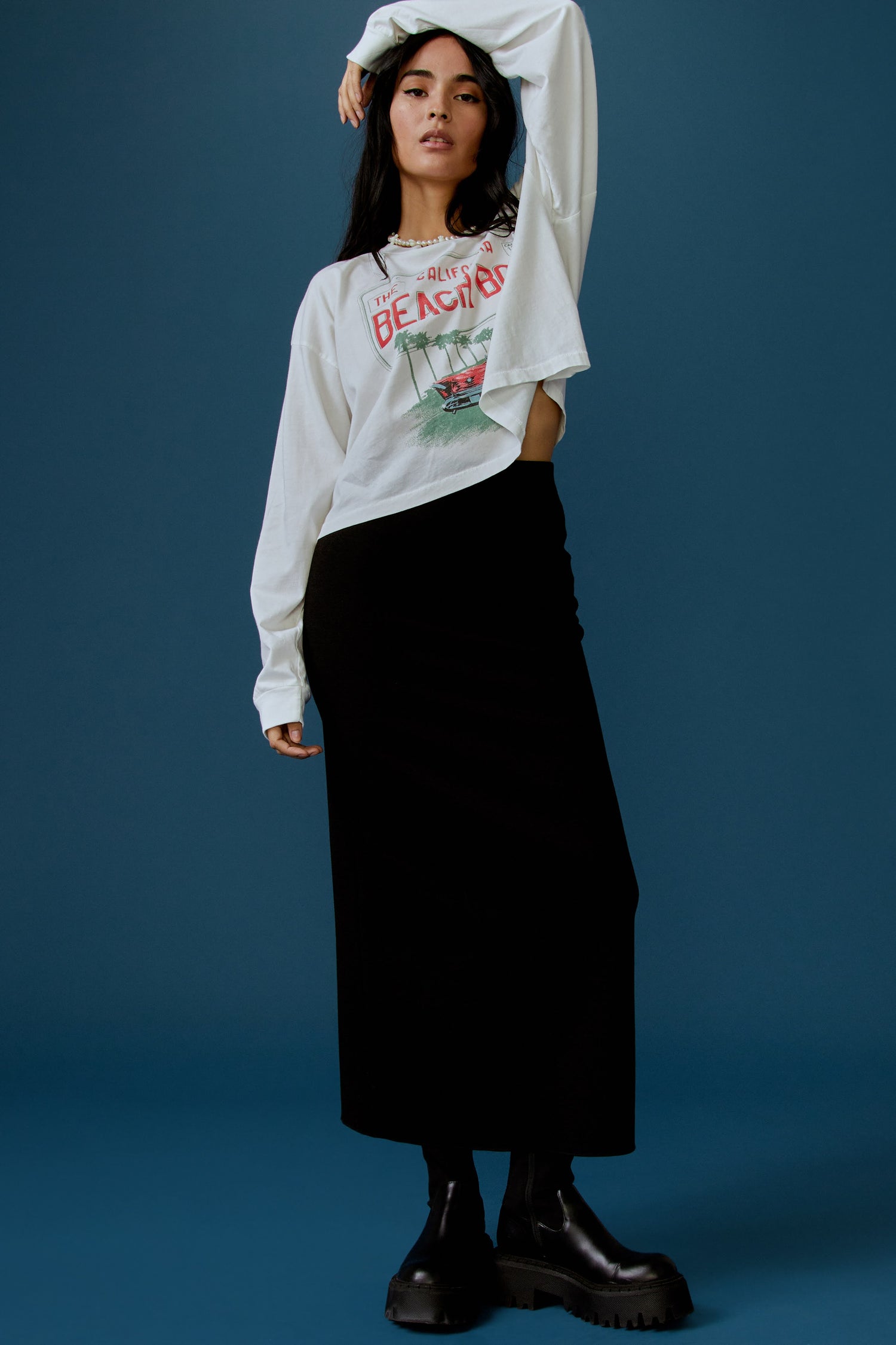 A dark-haired model featuring a white long sleeve stamped with "The Californa Beach Boys" and is desiged with a graphic of trees and cars on the center.
