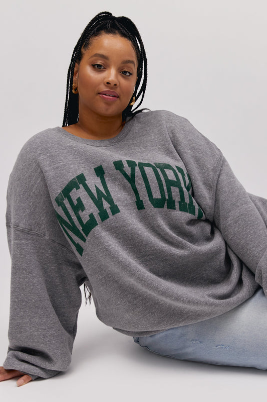 Plus size model wearing a grey crewneck sweatshirt with collegiate style 'New York' graphic lettering.