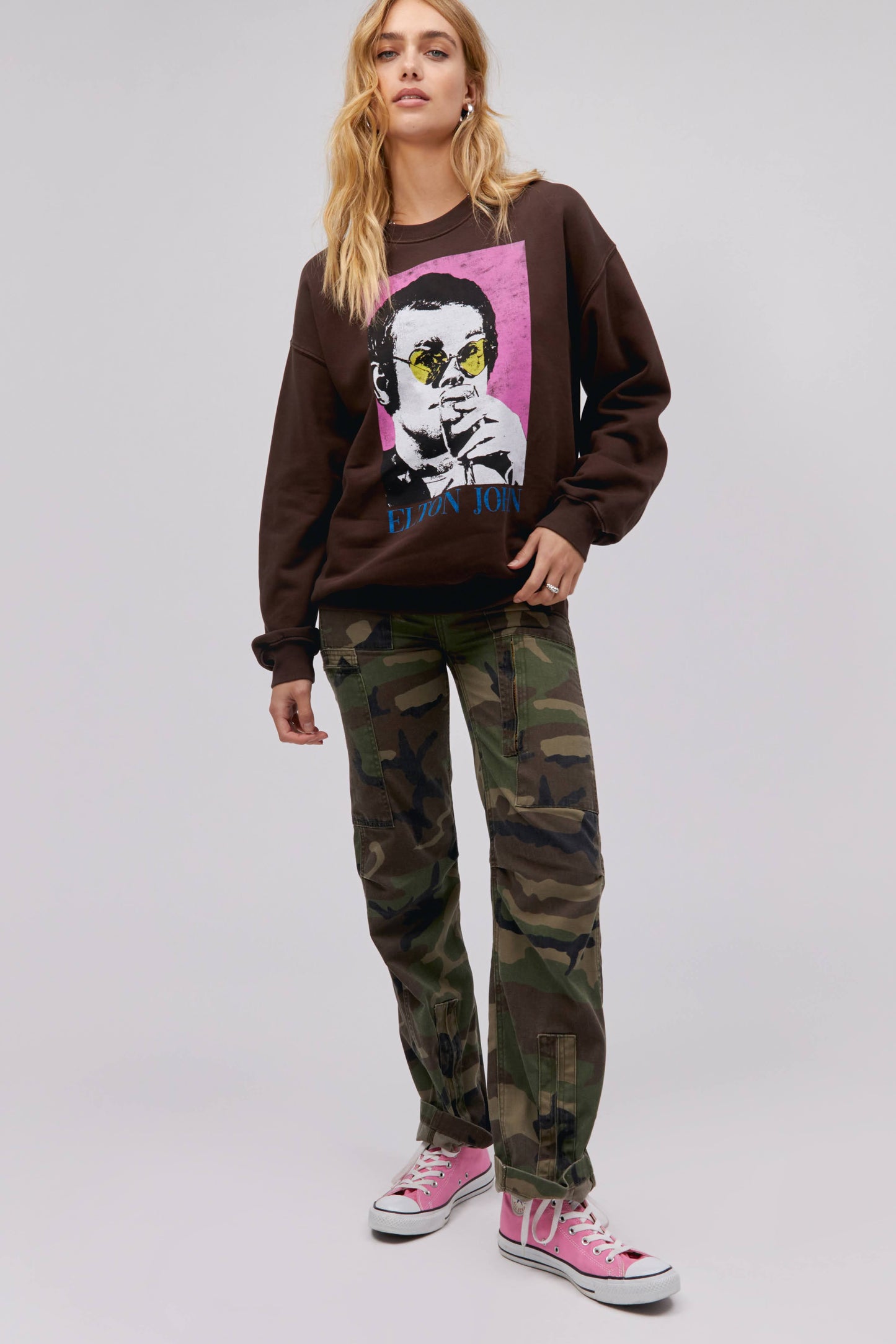 A blonde-haired model featuring a dark brown bf crew stamped with 'Elton John' and designed with an iconic portrait of the artist.