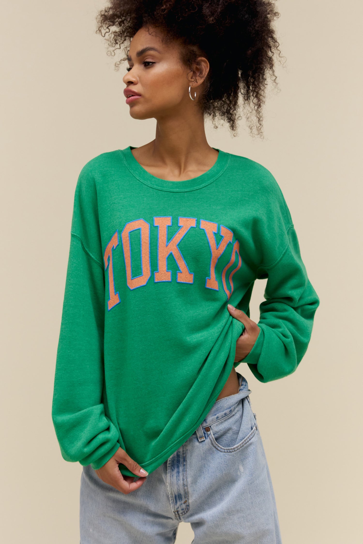 Curly-haired model wearing an oversized tri-blend fleece sweatshirt in green with contrast orange 'Tokyo' collegiate style lettering