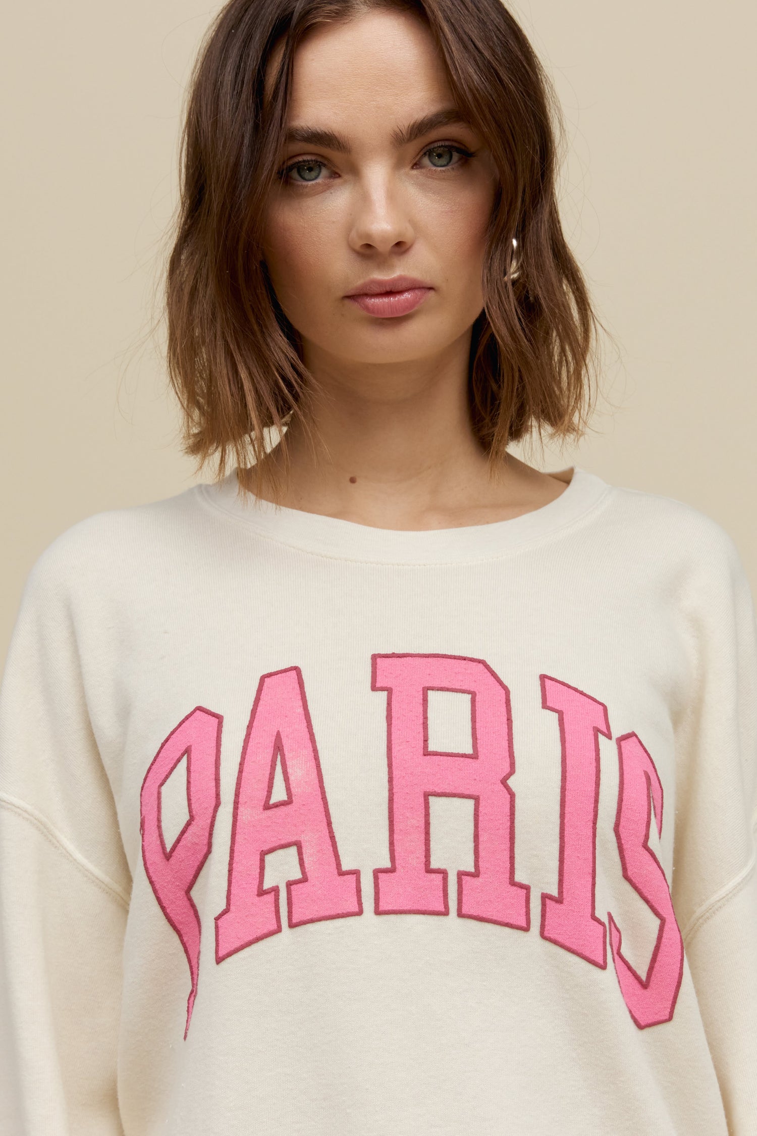 Short-haired model wearing an oversized tri-blend fleece sweatshirt in stone white with contrast pink 'Paris' collegiate style lettering