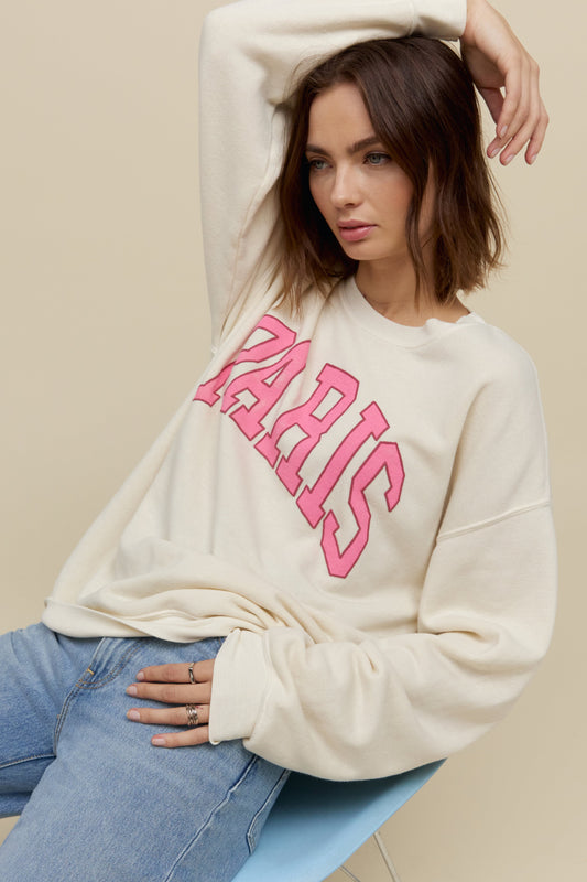 Short-haired model wearing an oversized tri-blend fleece sweatshirt in stone white with contrast pink 'Paris' collegiate style lettering