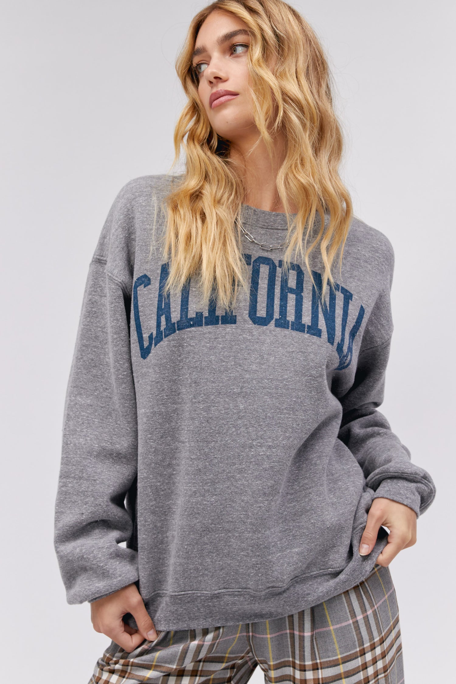 A blonde curly-haired model featuring a grey boyfriend crew stamped with 'California' in collegiate style letters.