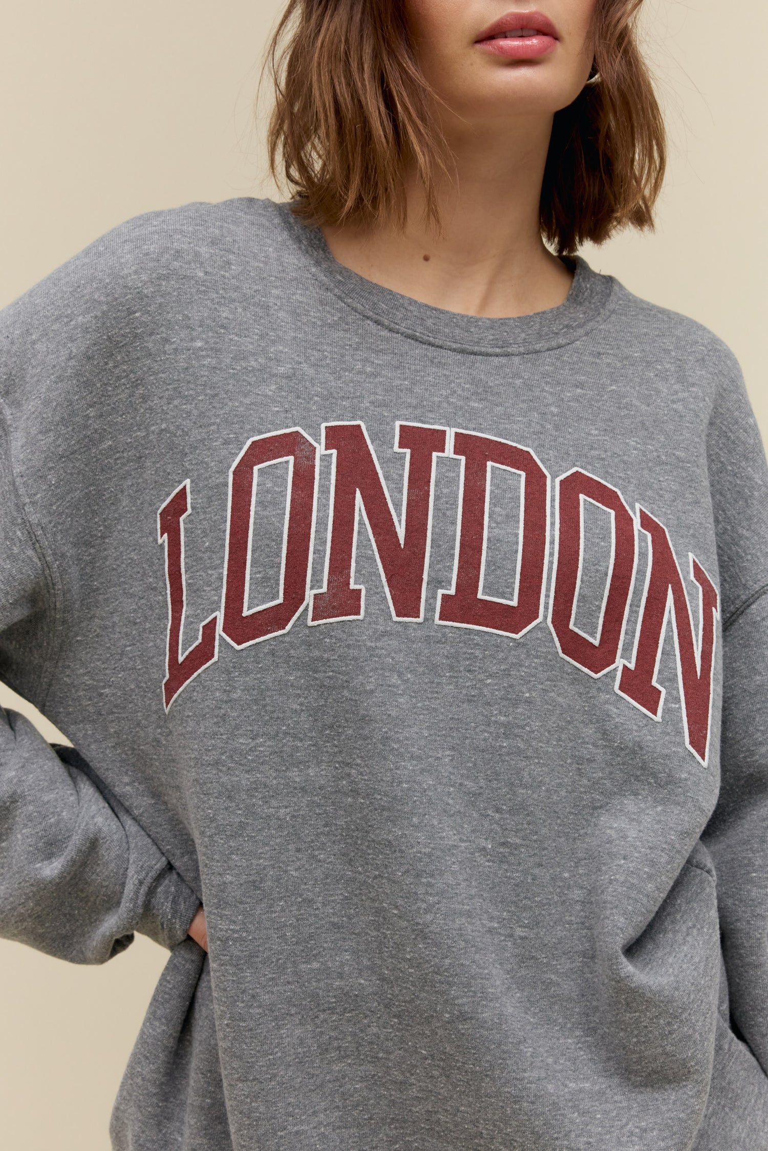 Short-haired model wearing an oversized tri-blend fleece sweatshirt in heather grey with contrast maroon 'London' collegiate style lettering