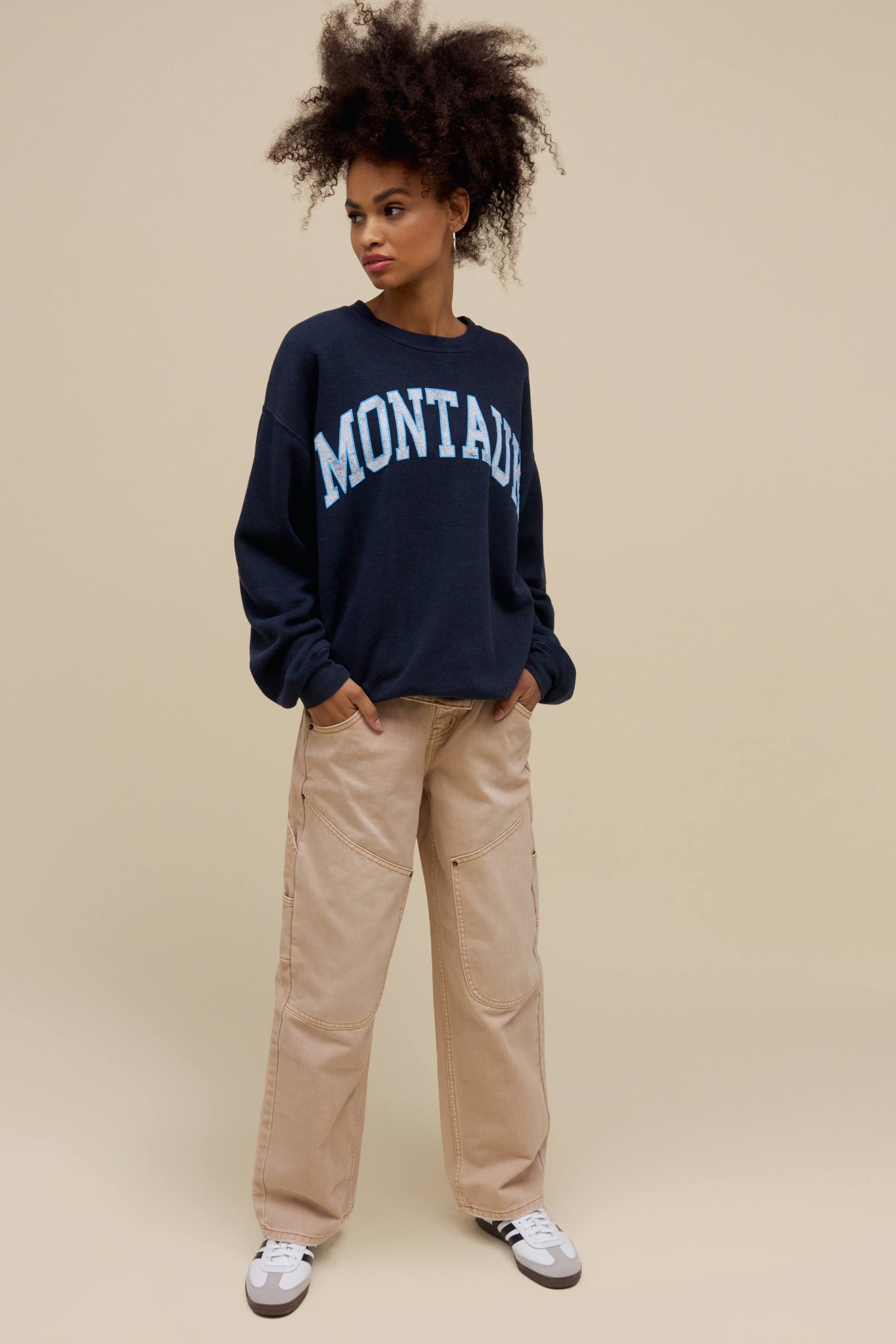 Curly-haired model wearing an oversized tri-blend fleece sweatshirt in navy heather with contrast light blue 'Montauk' collegiate style lettering