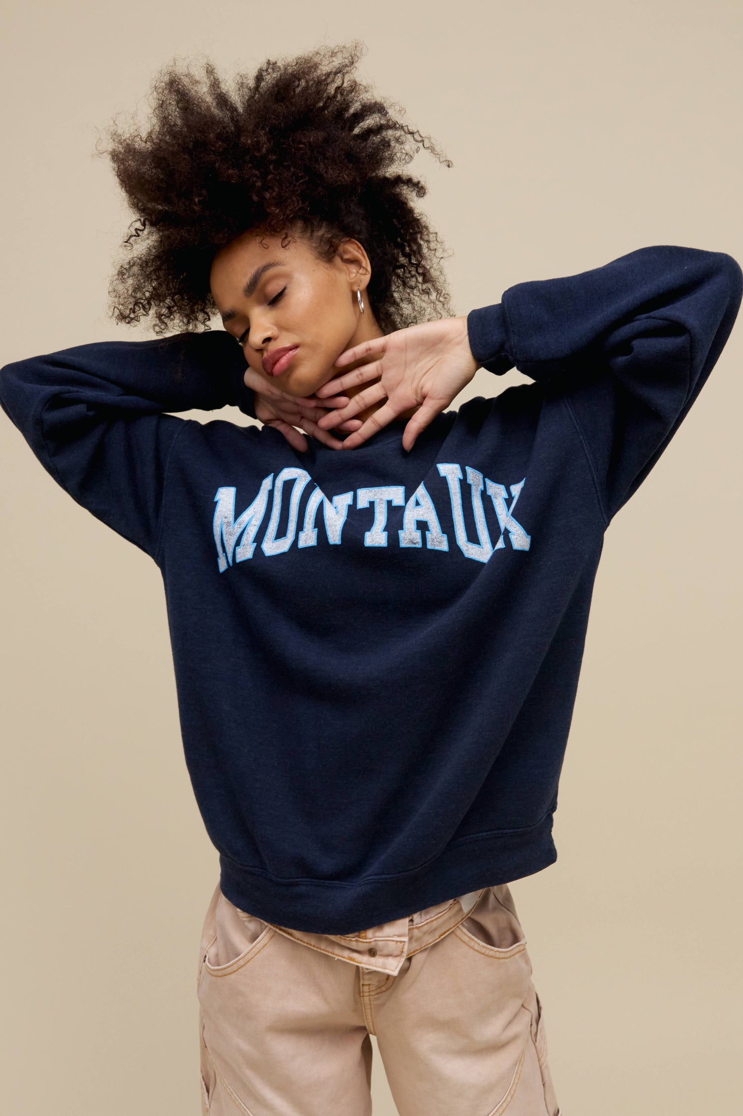 Curly-haired model wearing an oversized tri-blend fleece sweatshirt in navy heather with contrast light blue 'Montauk' collegiate style lettering
