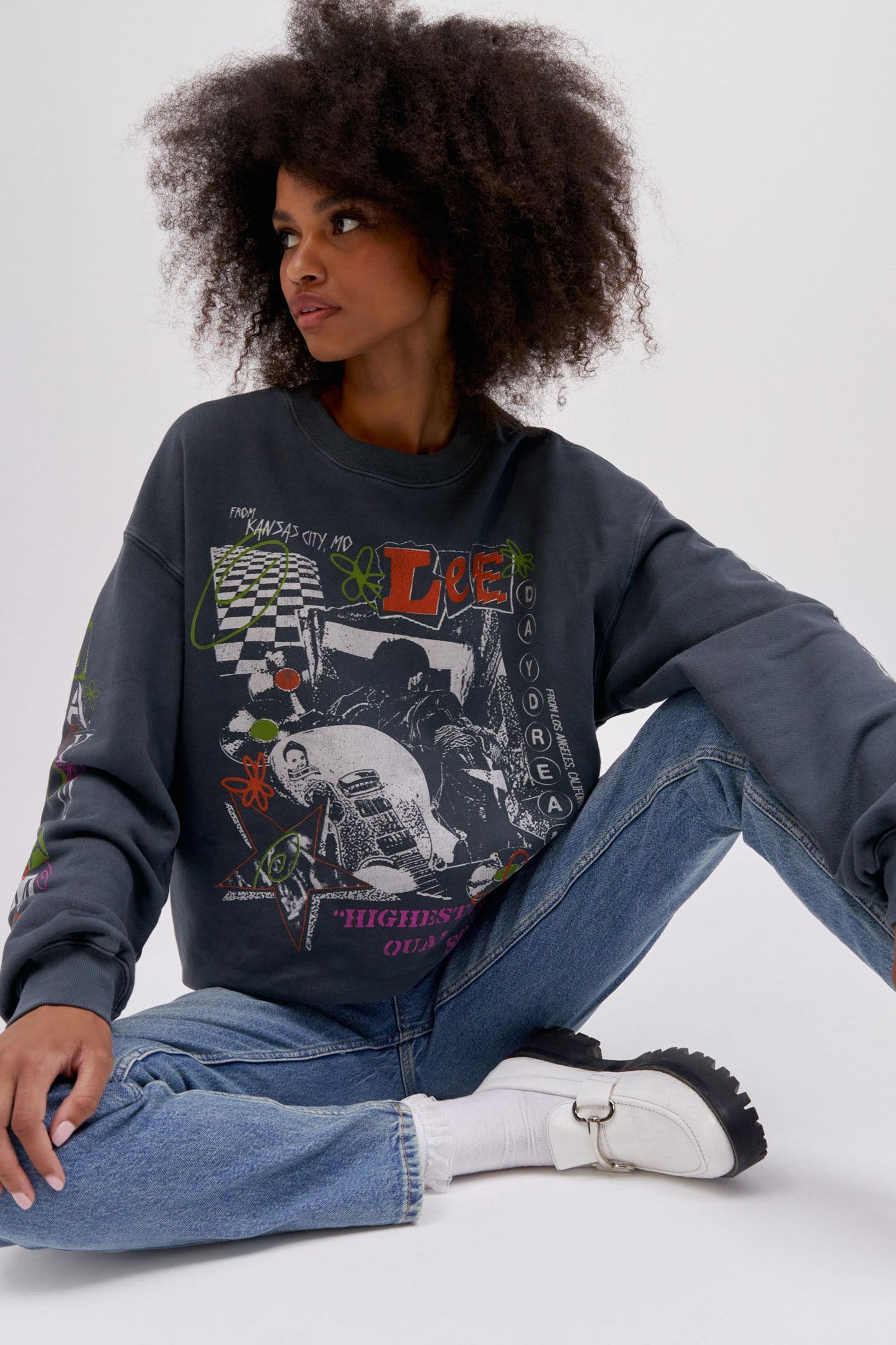 Sitting curly haired model wearing a Lee x Daydreamer collab graphic sweatshirt in washed black.