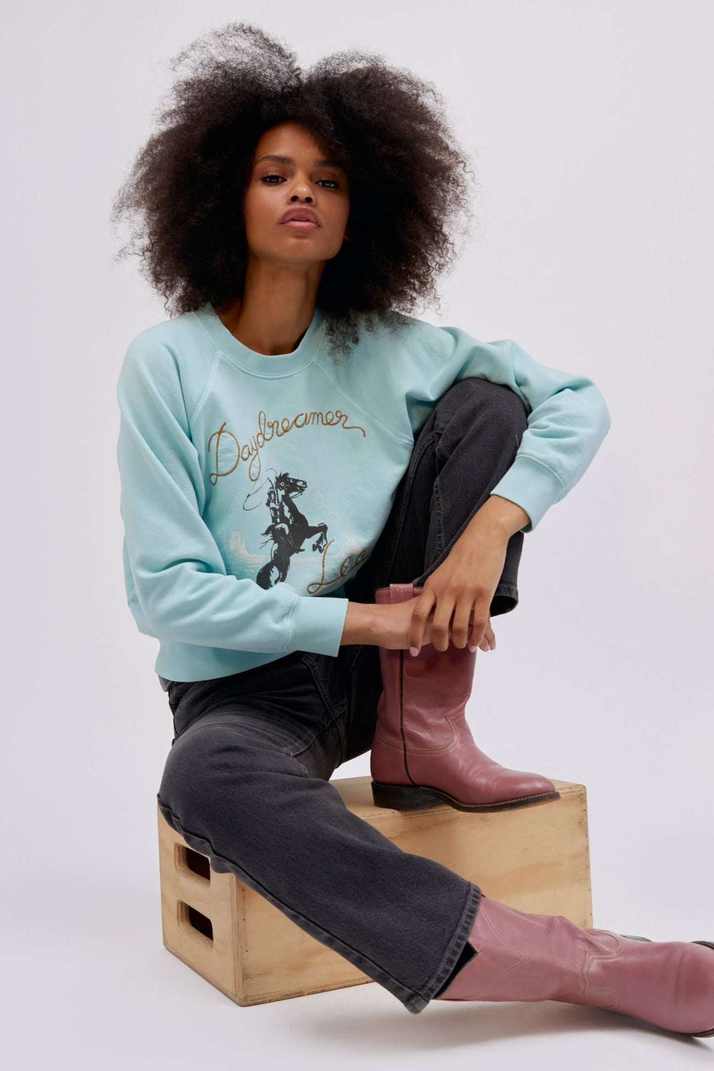A curly-haired model featuring a raglan crew in icy moon with a bucking cowgirl graphic inspired by the spirit of the West lands on a lightweight, french terry raglan crew embroidered with lasso lettering.