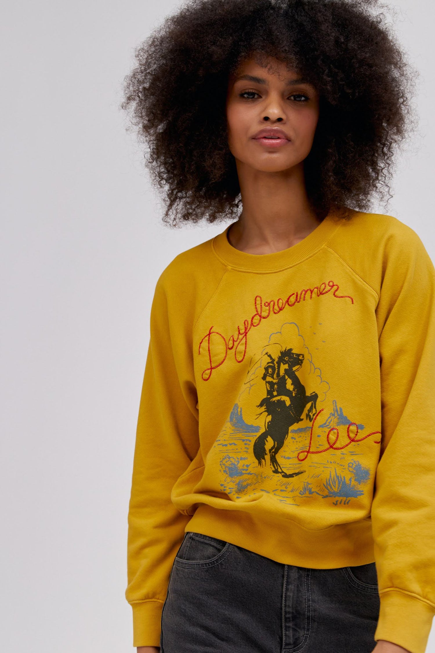 A curly-haired model featuring a raglan crew in golden daze with a bucking cowgirl graphic inspired by the spirit of the West lands on a lightweight, french terry raglan crew embroidered with lasso lettering.