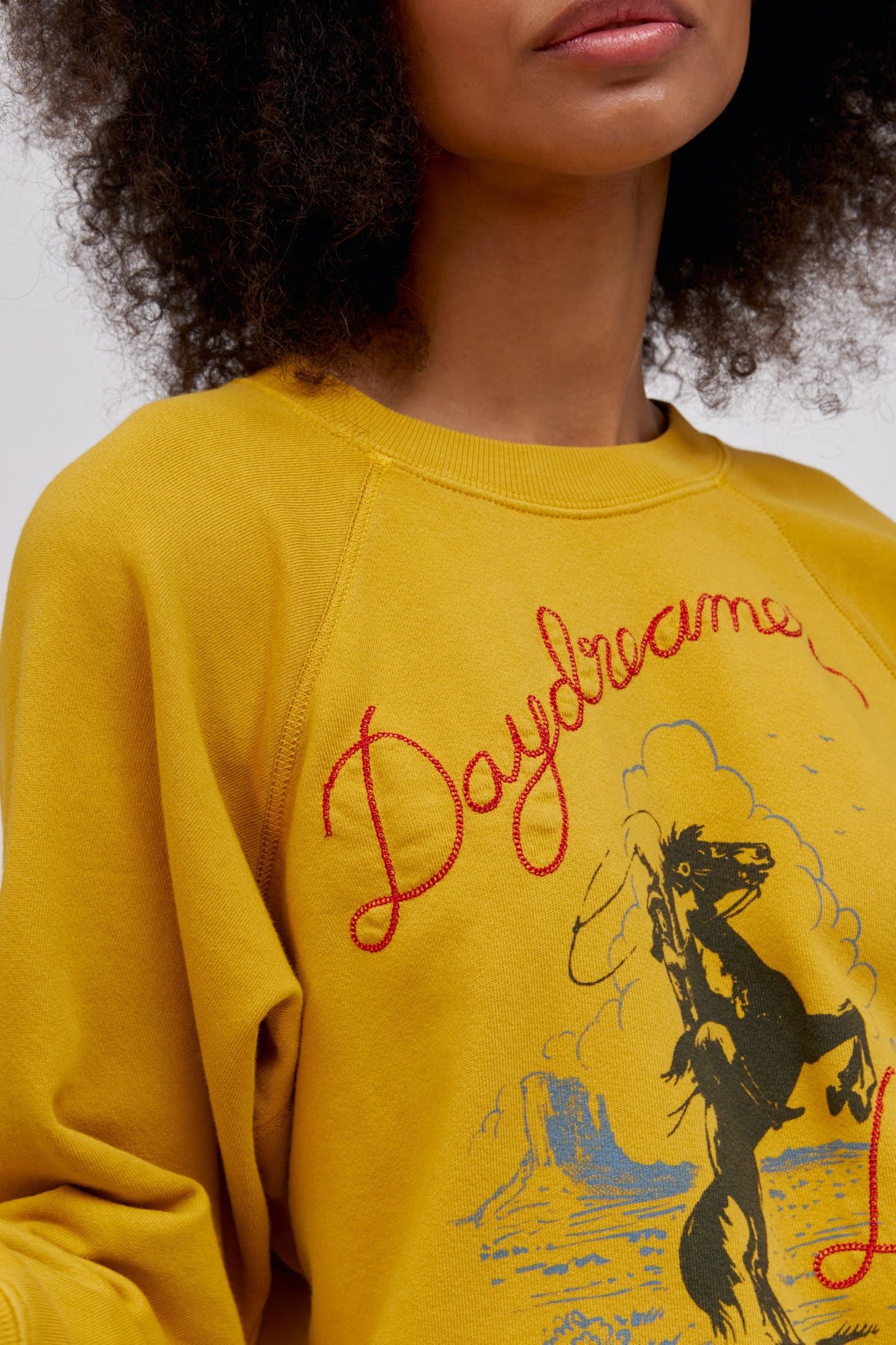 A curly-haired model featuring a raglan crew in golden daze with a bucking cowgirl graphic inspired by the spirit of the West lands on a lightweight, french terry raglan crew embroidered with lasso lettering.