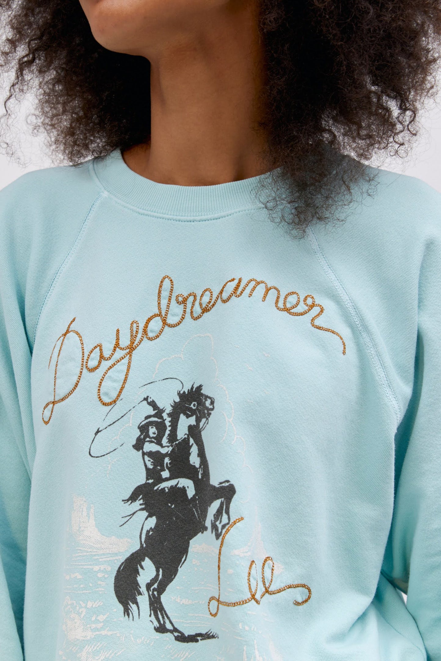 A curly-haired model featuring a raglan crew in icy moon with a bucking cowgirl graphic inspired by the spirit of the West lands on a lightweight, french terry raglan crew embroidered with lasso lettering.