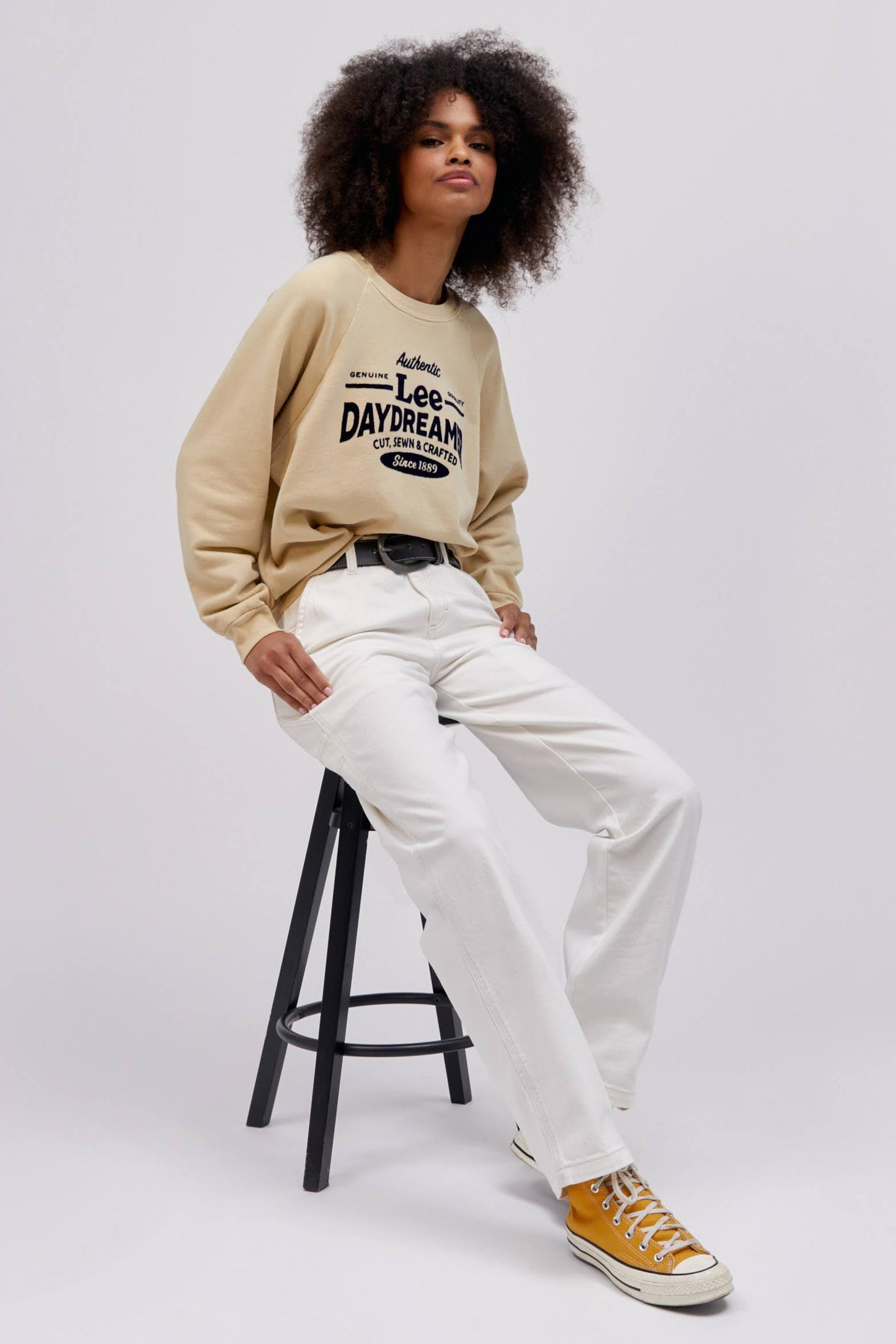 curly haired model sitting on stool wearing khaki colored sweatshirt and white workwear pants