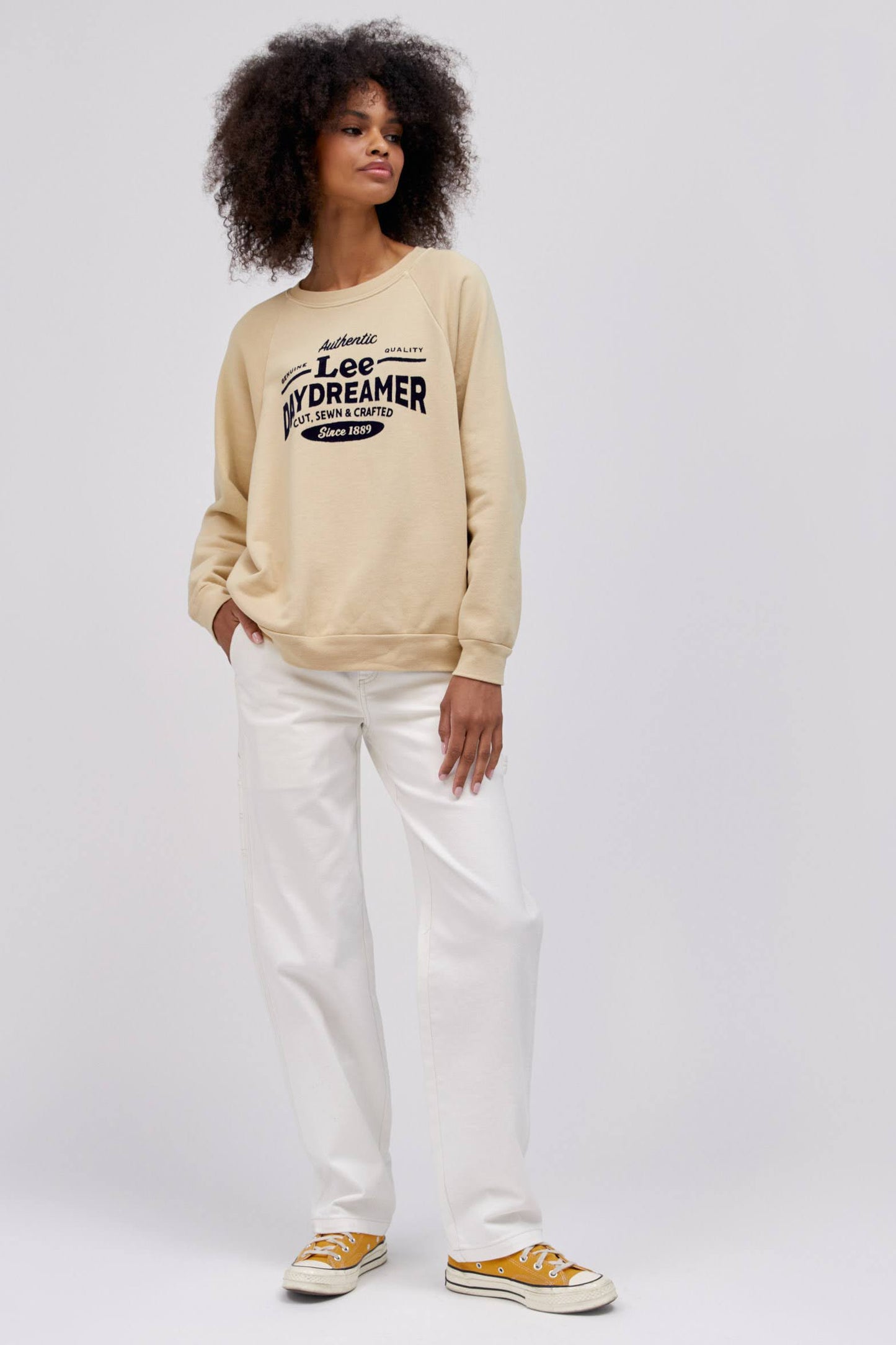 curly haired model wearing khaki colored sweatshirt and posing with hand in pocket of white workwear pants