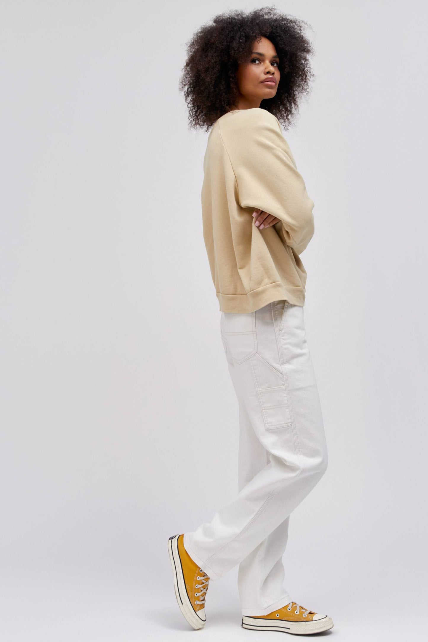 curly haired model in sideways arm crossed pose wearing khaki colored sweatshirt and white workwear pants