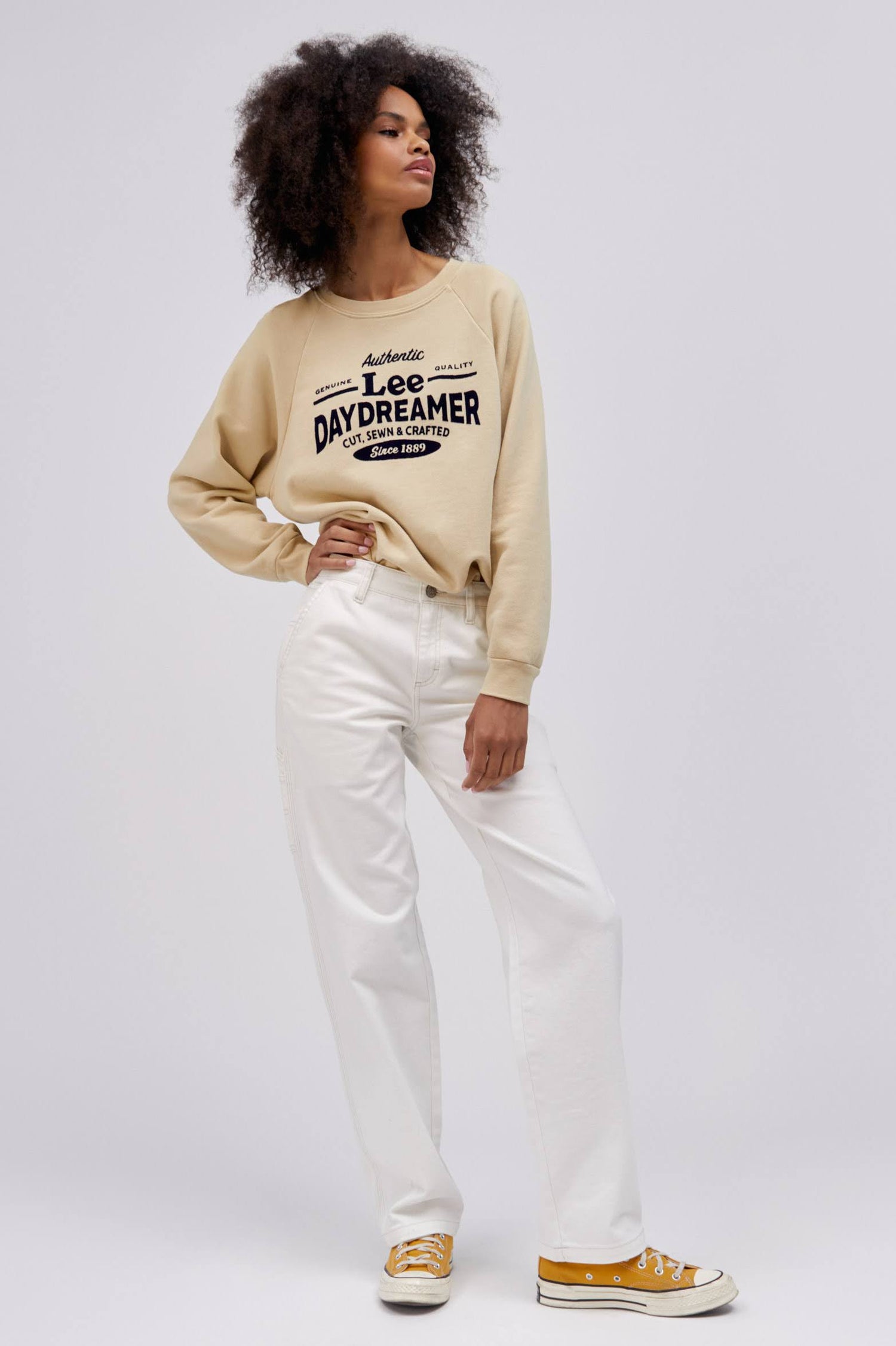 curly haired model posing with hand on hip wearing khaki colored sweatshirt and white workwear pants