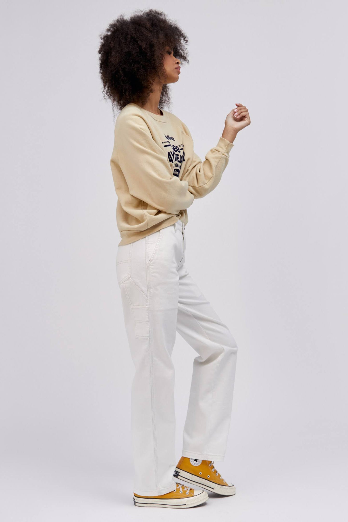 curly haired model posing to the side with hand up and wearing a khaki sweatshirt and white workwear pants