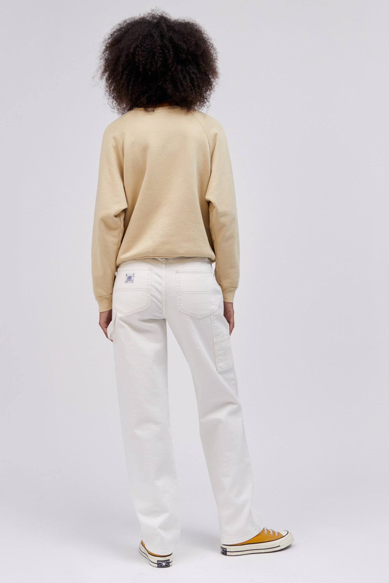 backside of curly haired model in a standing pose wearing a khaki colored sweatshirt and white workwear pants